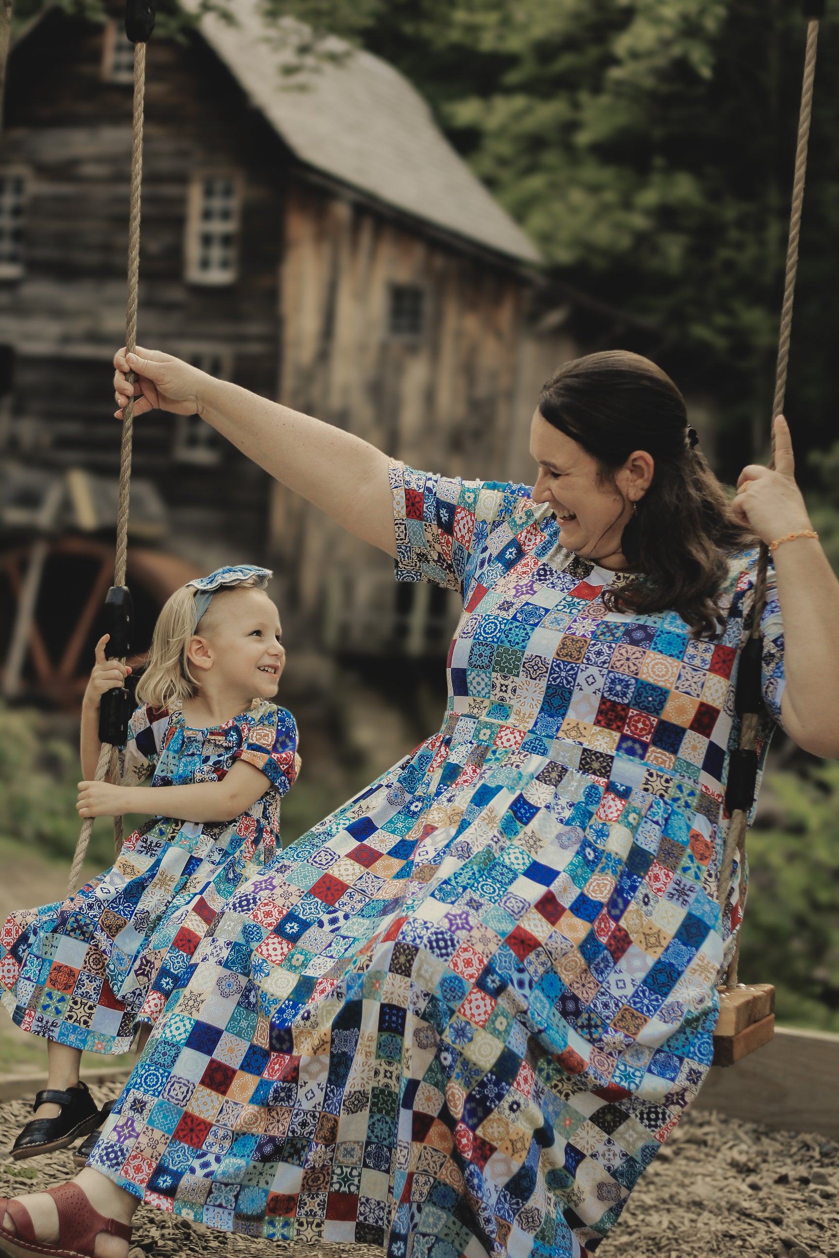 Mother and daughter in colorful patchwork modest dresses on a swing set with a rustic wooden cabin in the background.