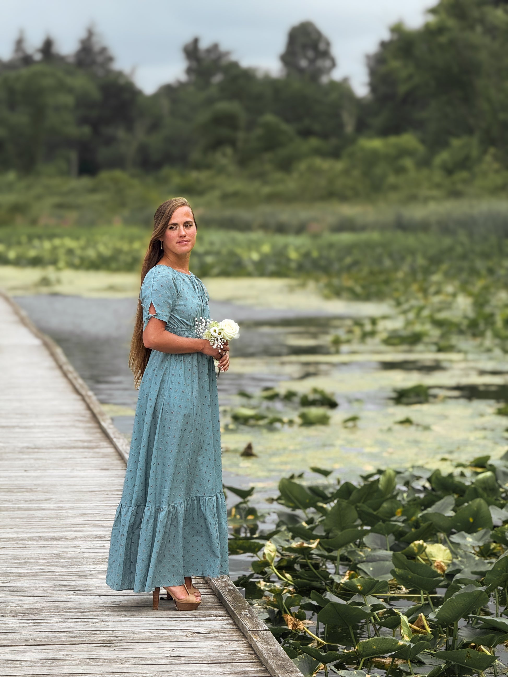 Woman in a blue modest nursing dress standing on a wooden dock by a body of water with greenery.