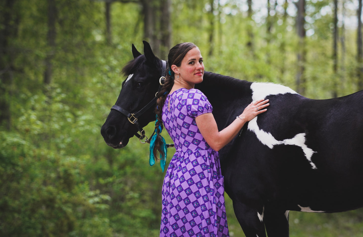 Woman in a purple modest nursing dress standing with a horse in a forested area