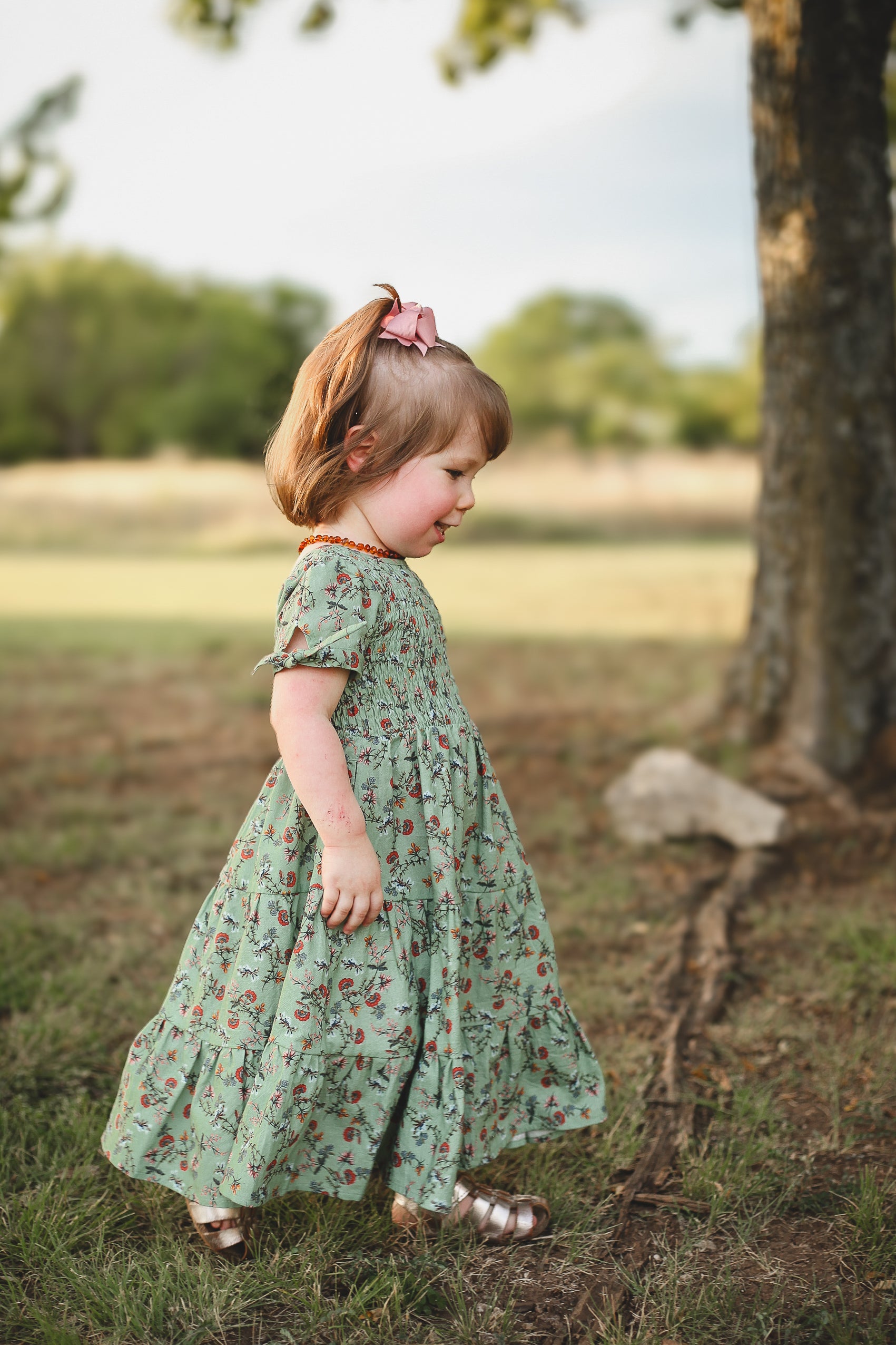 Young girl in a green floral dress standing in a natural setting with trees and grass.
