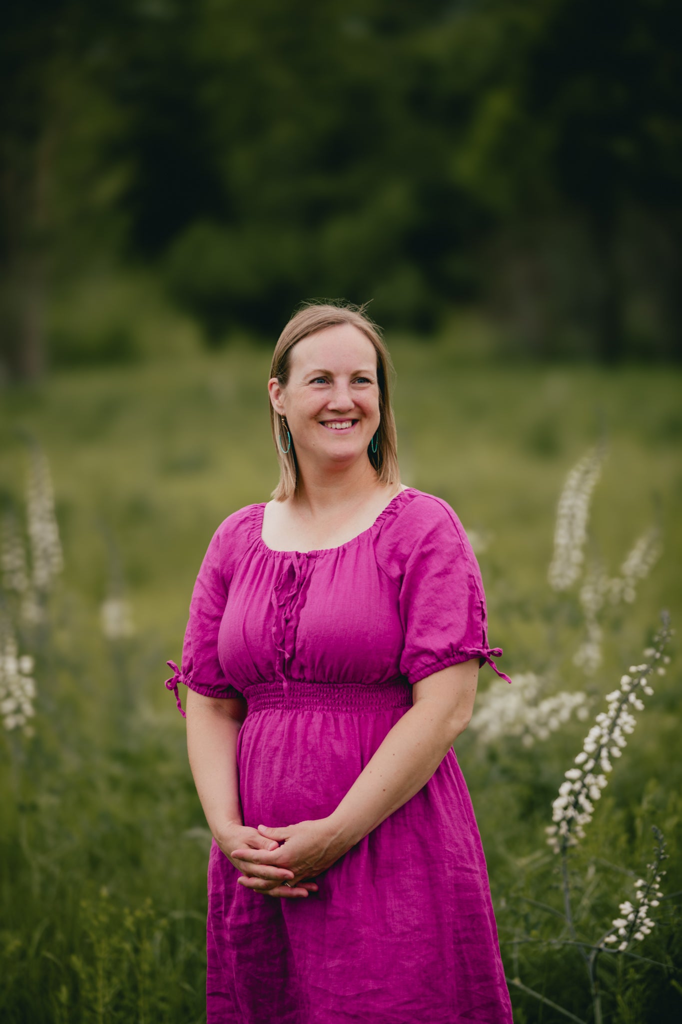 Woman in a pink modest nursing dress standing in a field with greenery and white flowers.