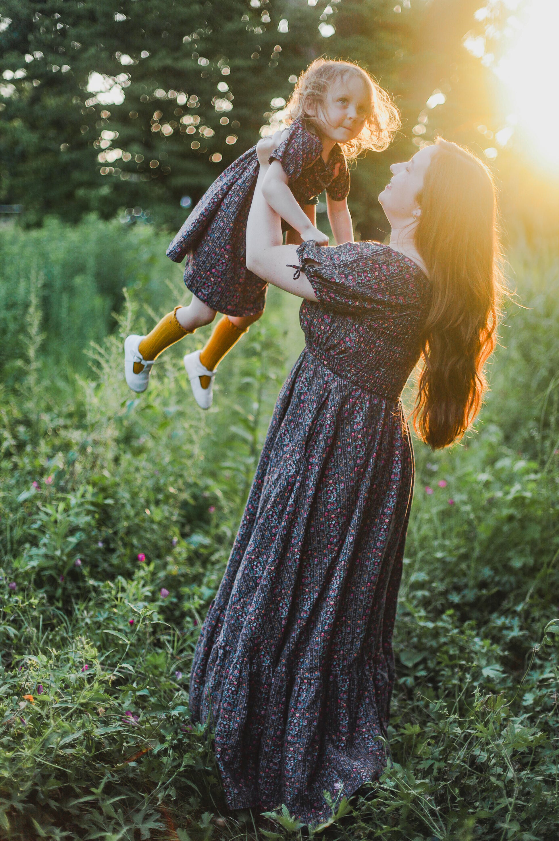 Woman holding a child in a field with sunlight filtering through wearing a modest nursing dress
