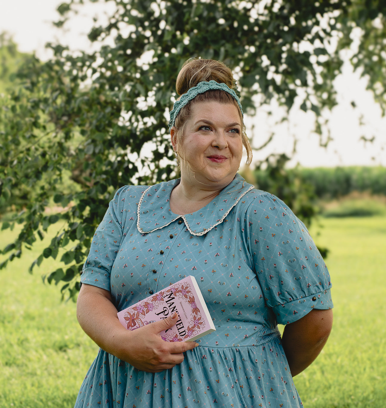 Woman in a blue modest nursing dress holding a book in a green outdoor setting