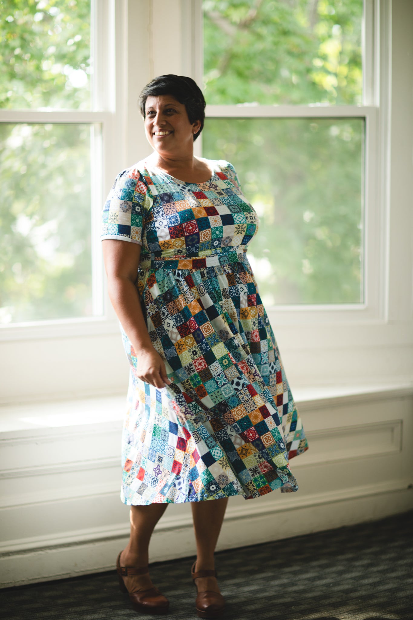 Woman wearing a colorful patchwork modest nursing dress standing in front of a window with greenery outside.