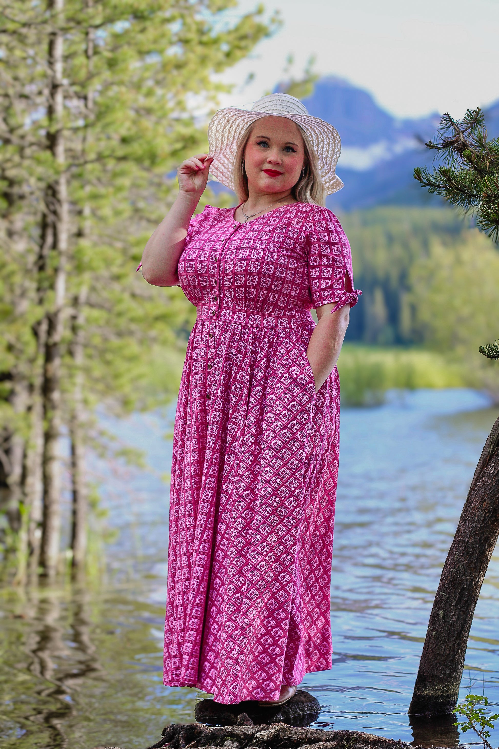 Woman in a pink modest nursing dress and hat standing by a lake with mountains in the background