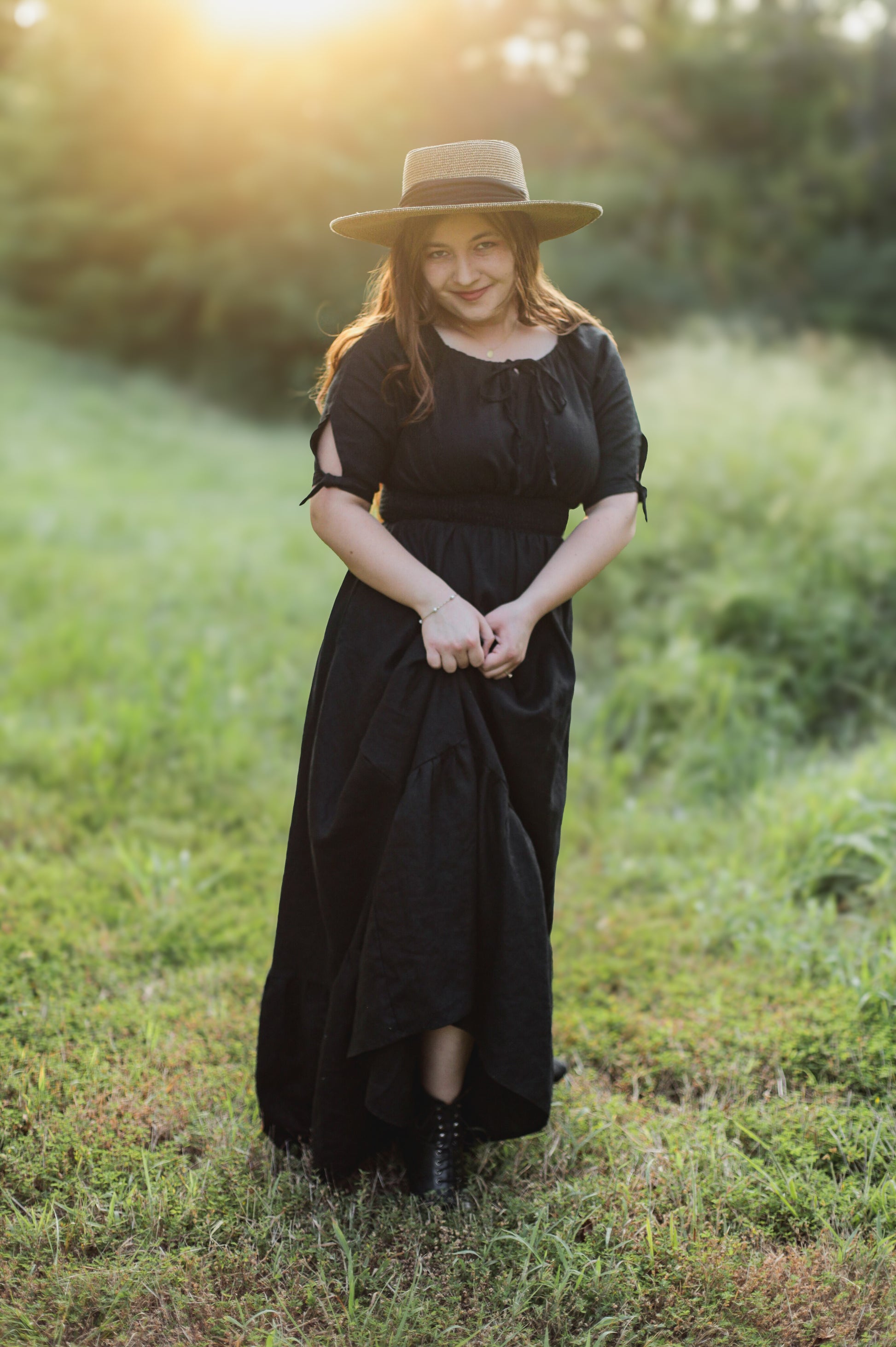 Woman in a black modest nursing dress and hat standing in a grassy field with sunlight filtering through.
