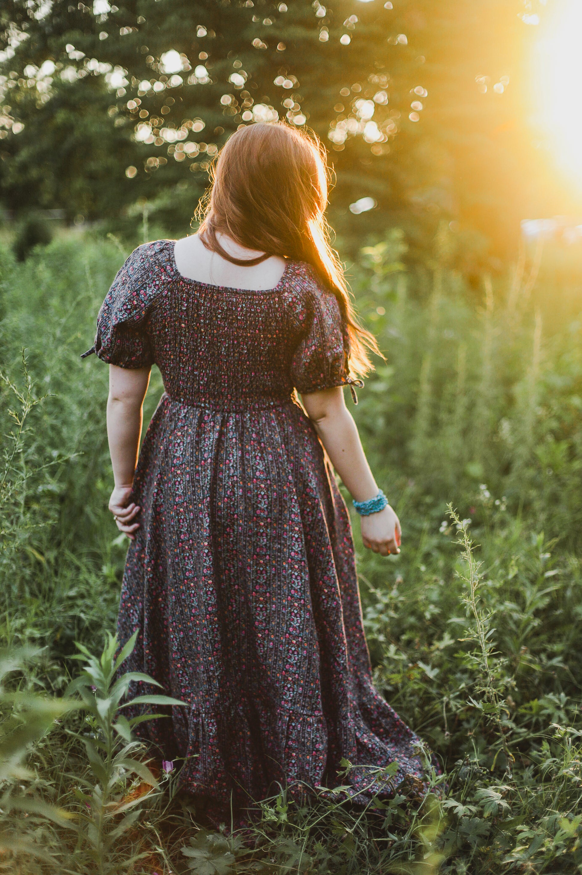 Woman in a long modest nursing dress standing in a field with sunlight filtering through the trees