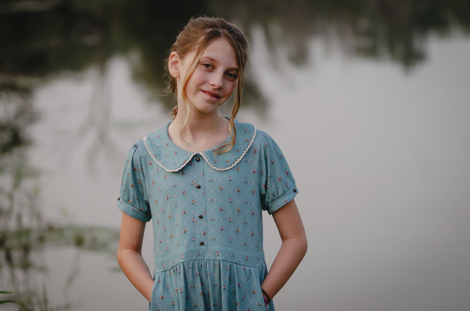 Young girl in a modest blue dress standing by a body of water with a blurred background