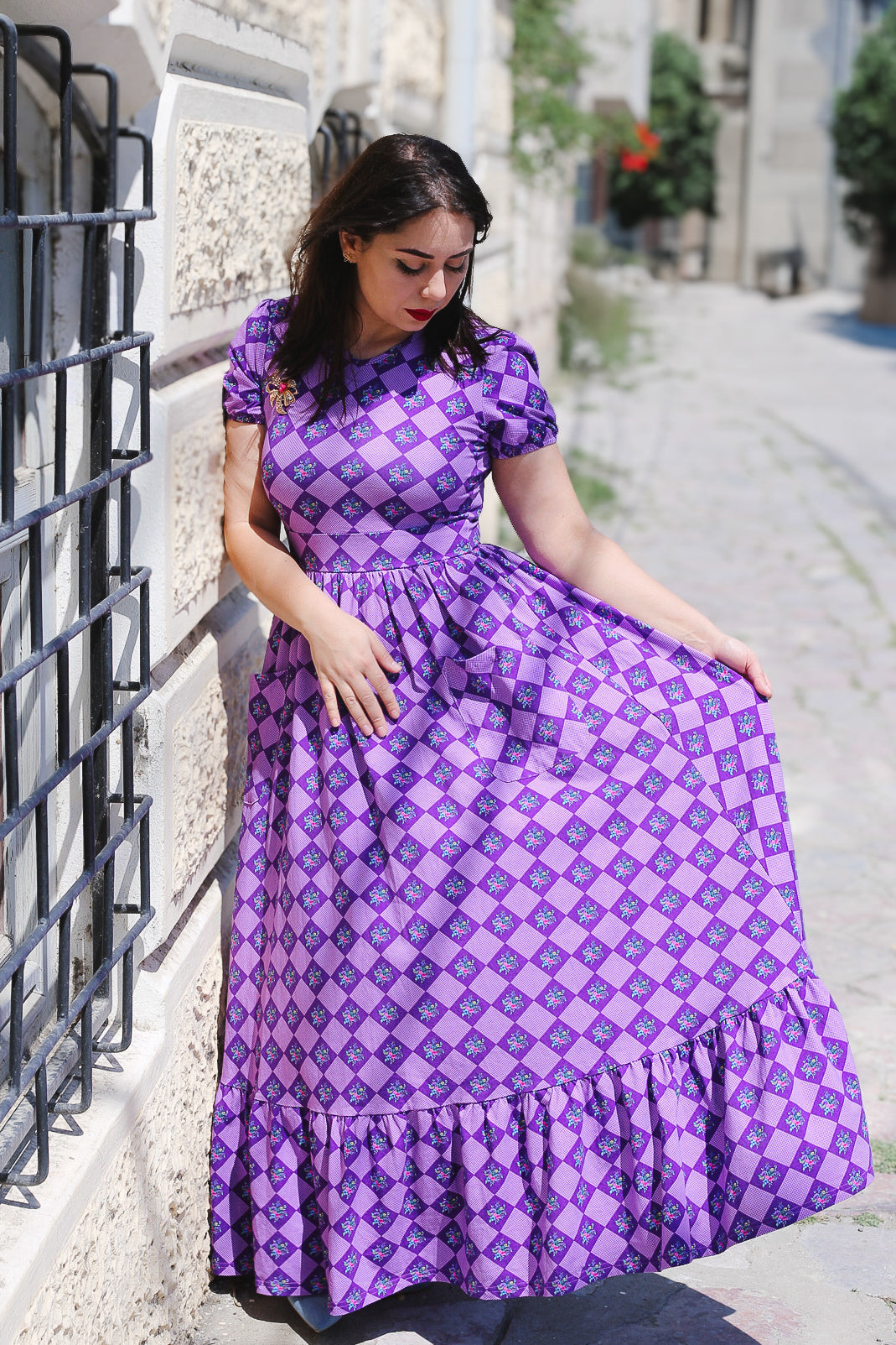 Woman in a purple checkered modest nursing dress leaning against a wall on a street.
