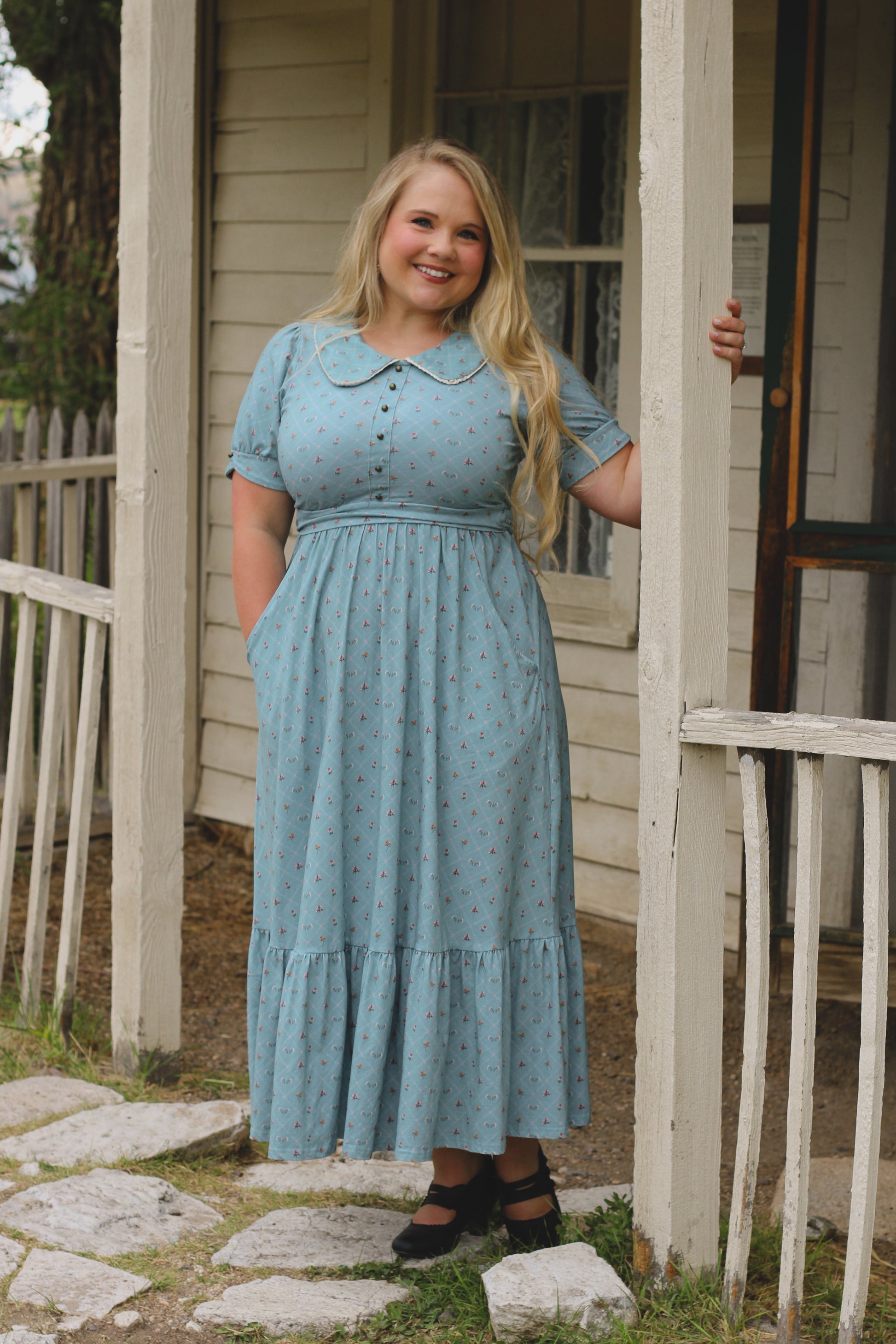 Woman in a light blue modest nursing dress standing outside a house.