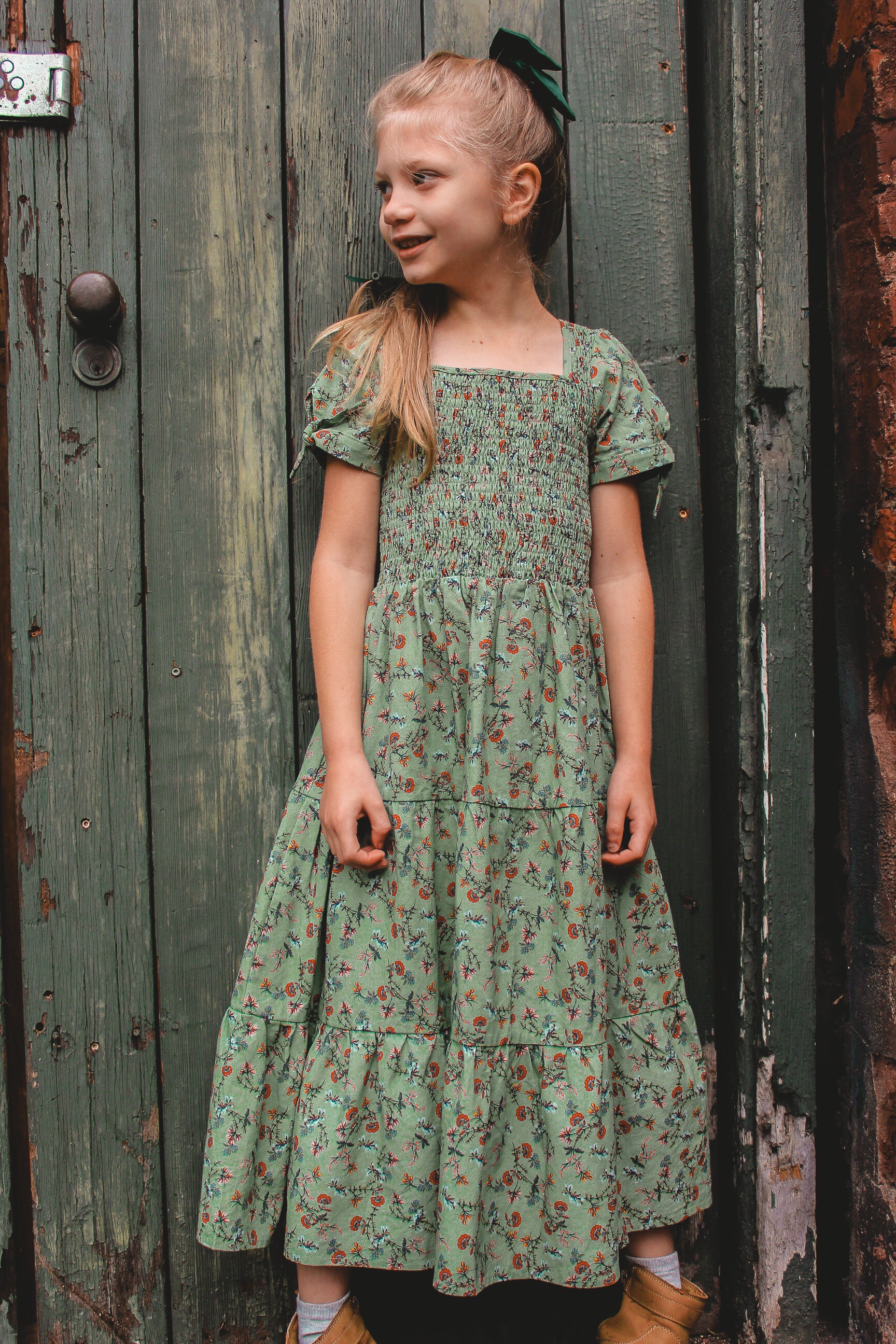 Young girl in a green floral dress standing against a rustic wooden door.
