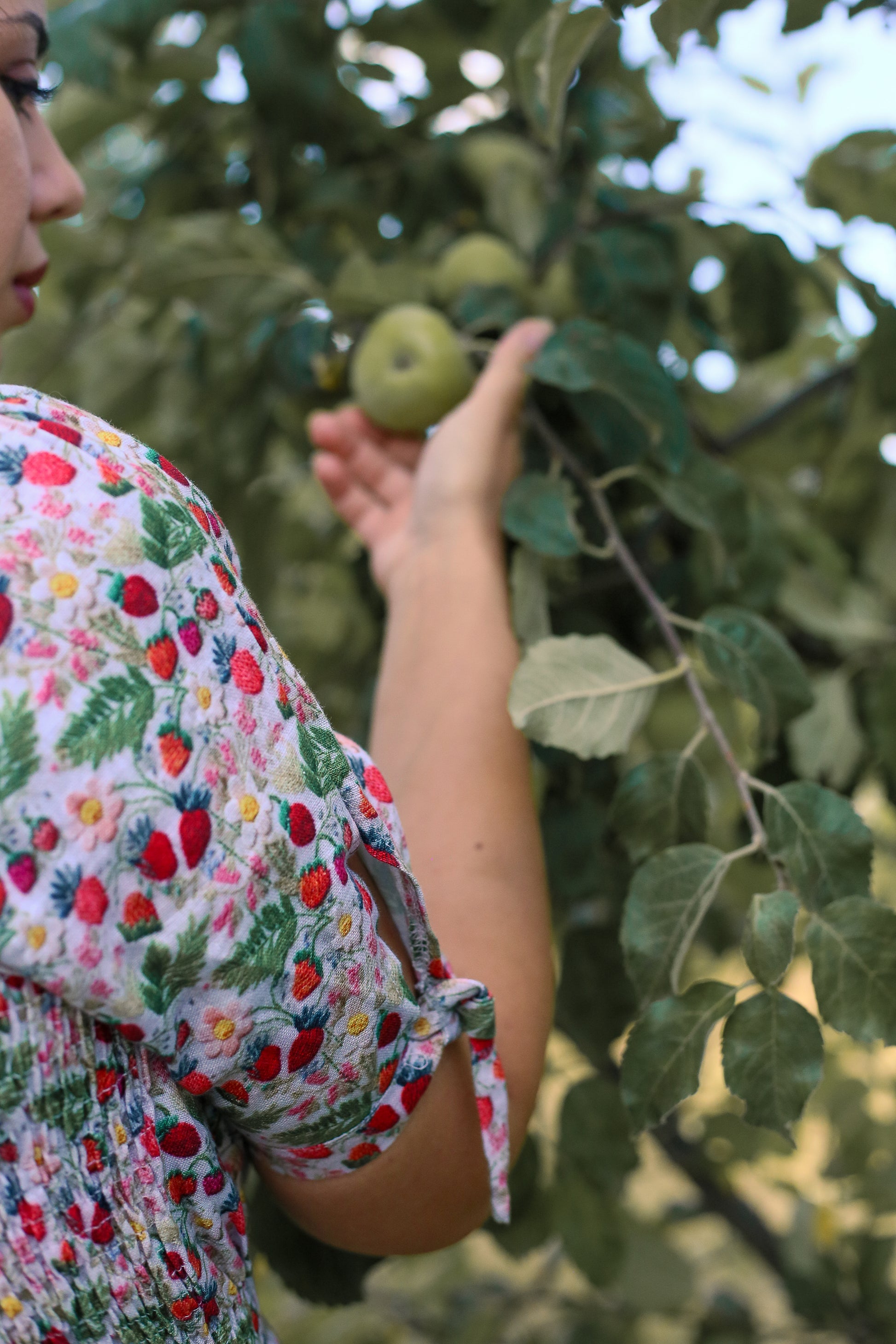Person in a floral modest nursing dress reaching out to touch an apple on a tree