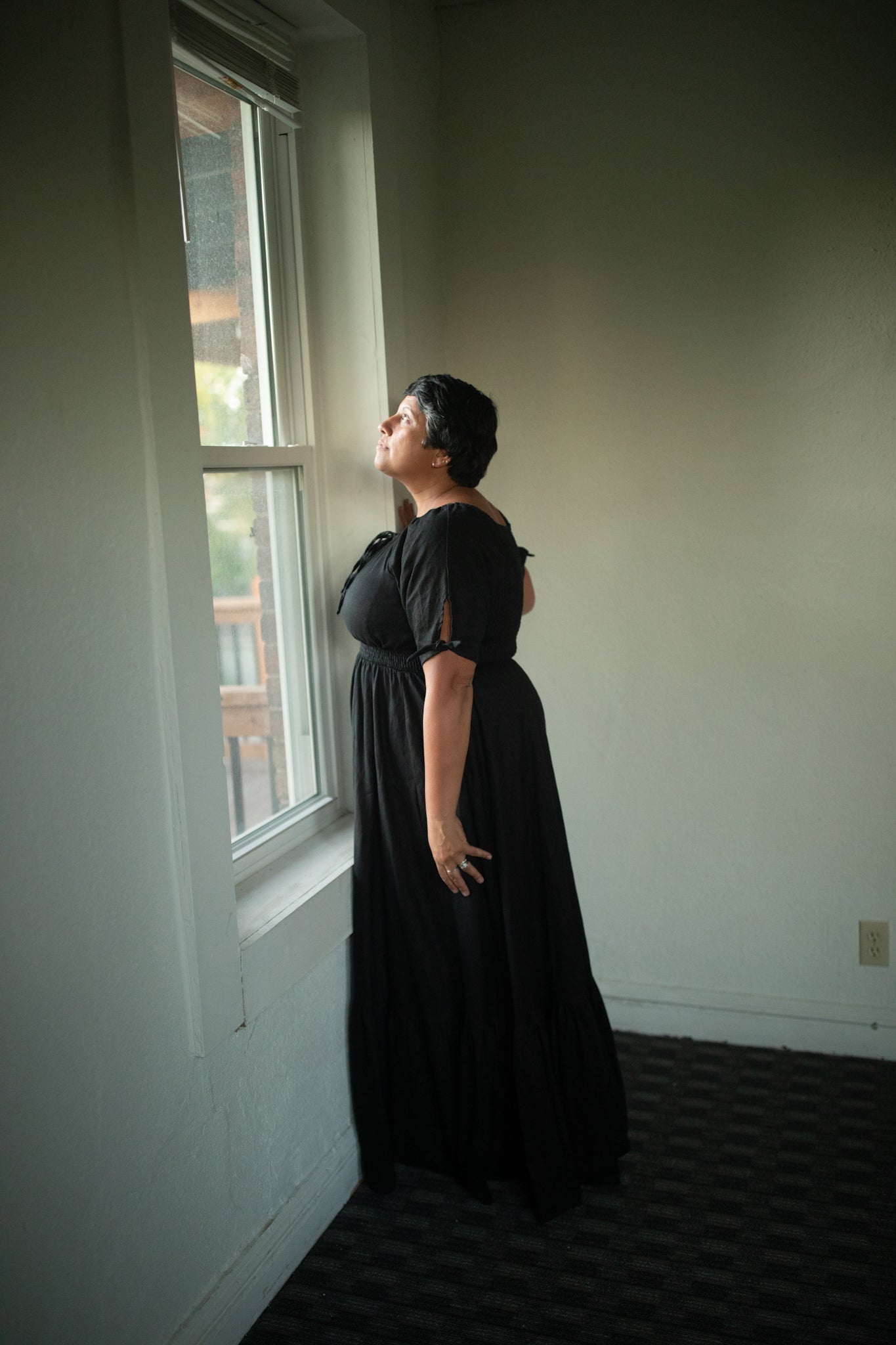 Woman in a black modest nursing dress standing by a window in a room with plain walls.