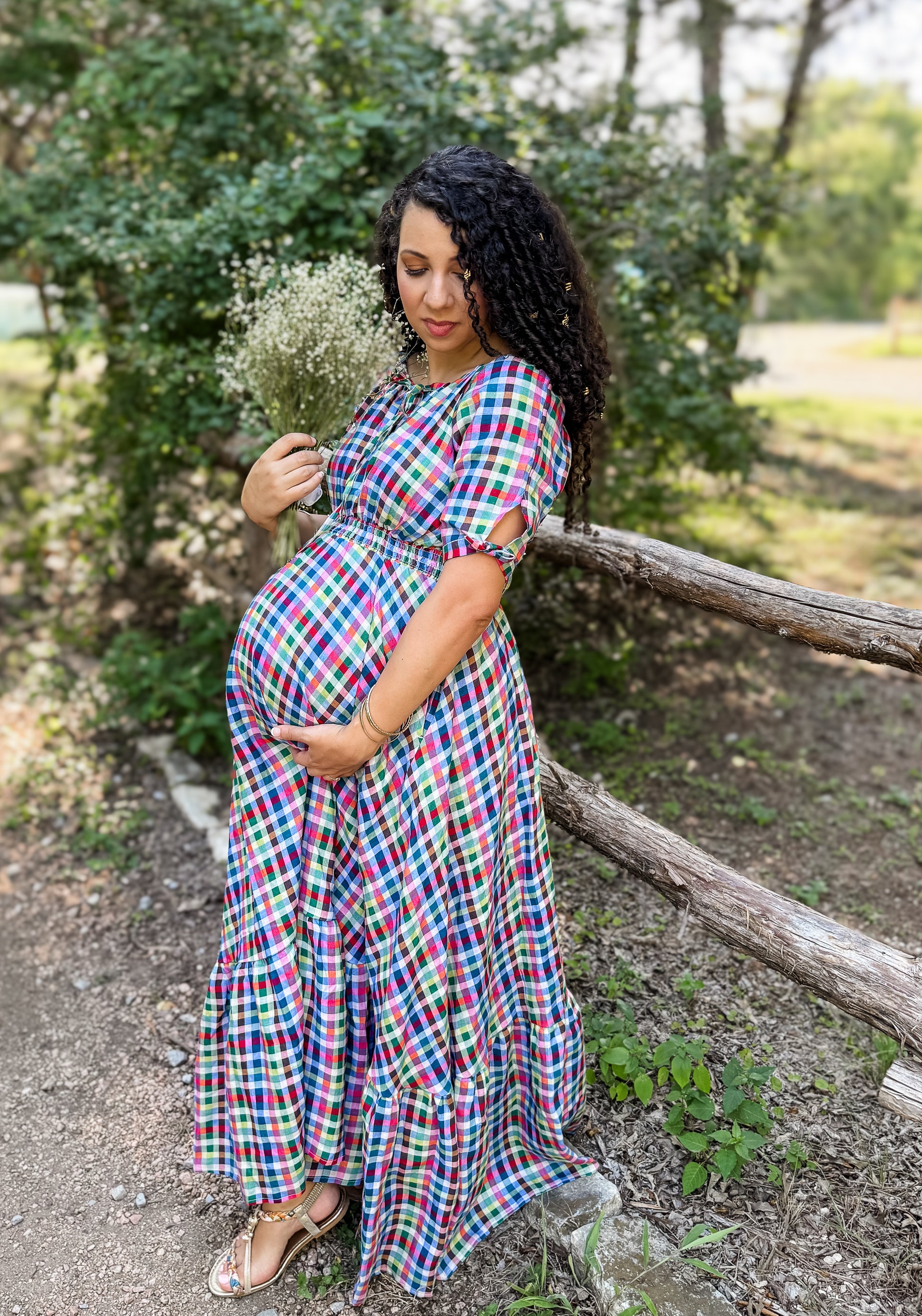 Pregnant woman in a colorful plaid modest nursing dress standing outdoors near a wooden fence.