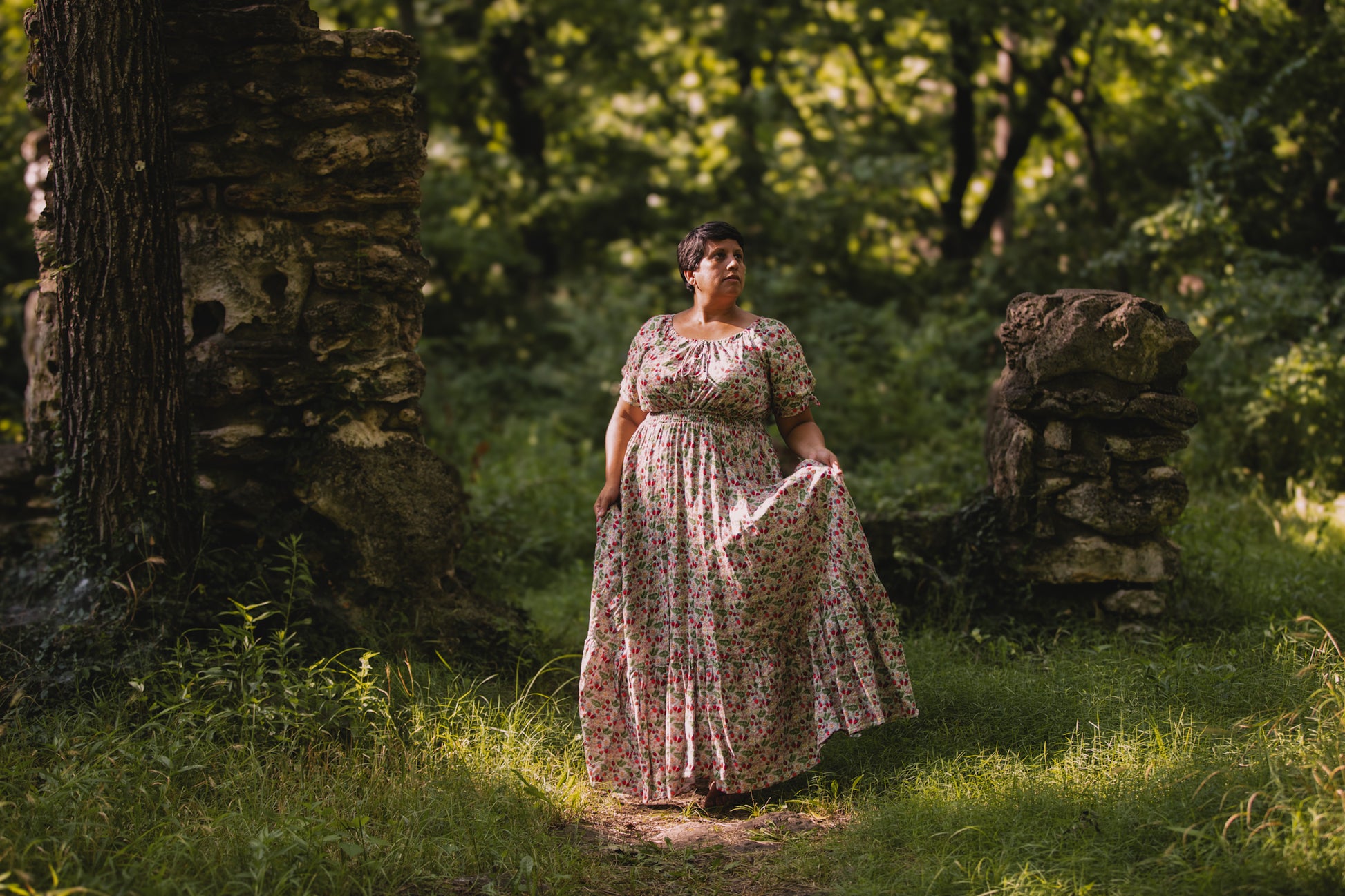 Woman in a floral modest nursing dress standing in a forested area with sunlight filtering through the trees.