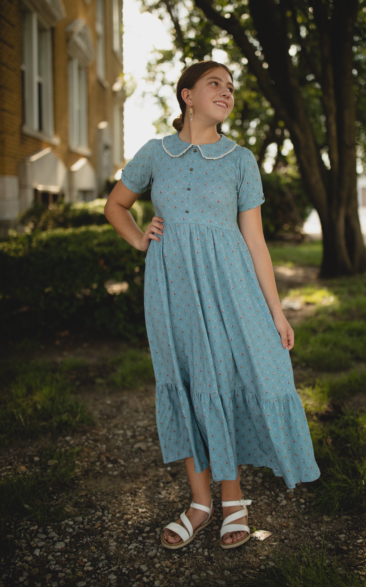 Woman in a blue modest nursing dress standing outdoors with trees and a building in the background