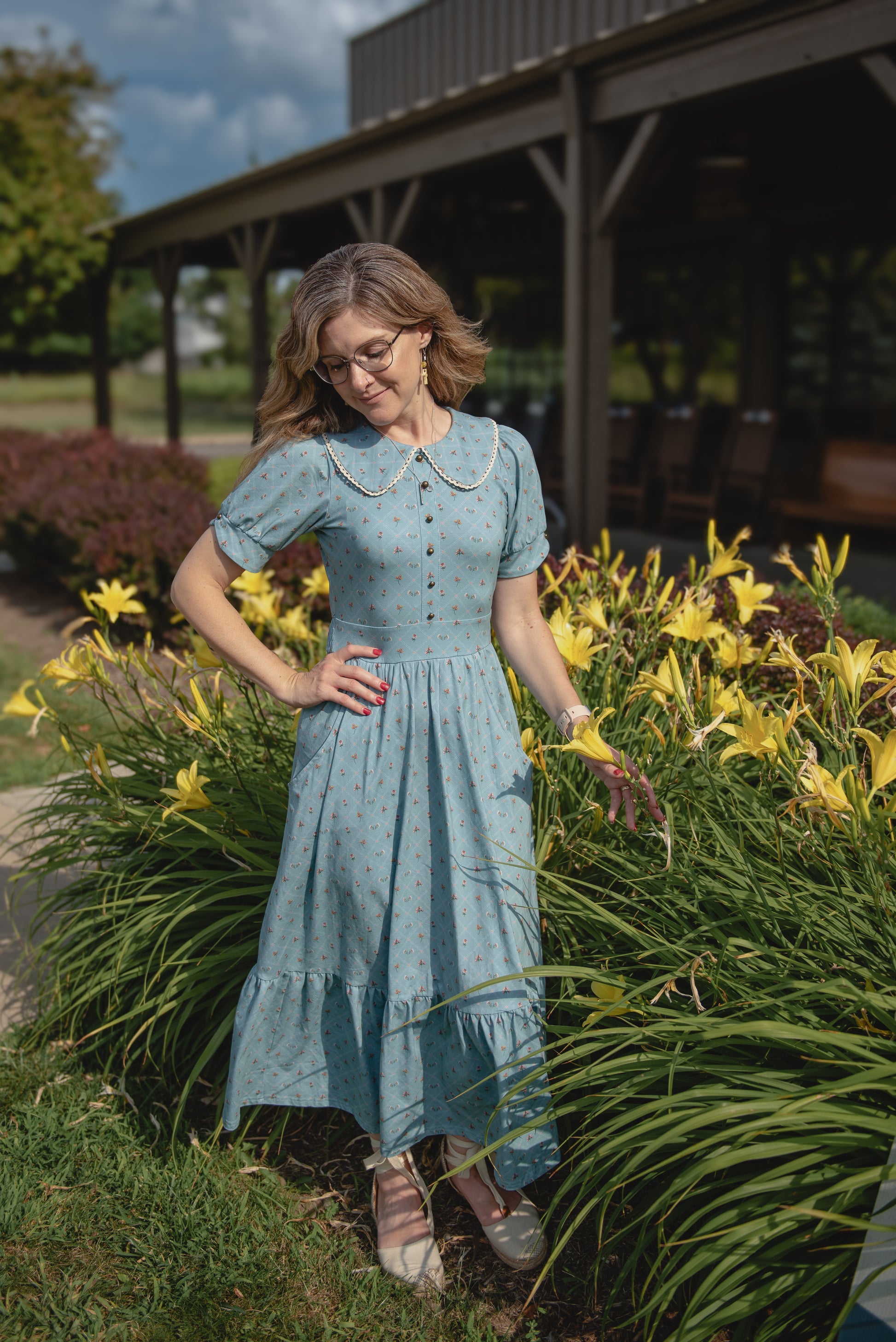 Woman in a light blue modest nursing dress standing in a garden with yellow flowers and a wooden structure in the background.