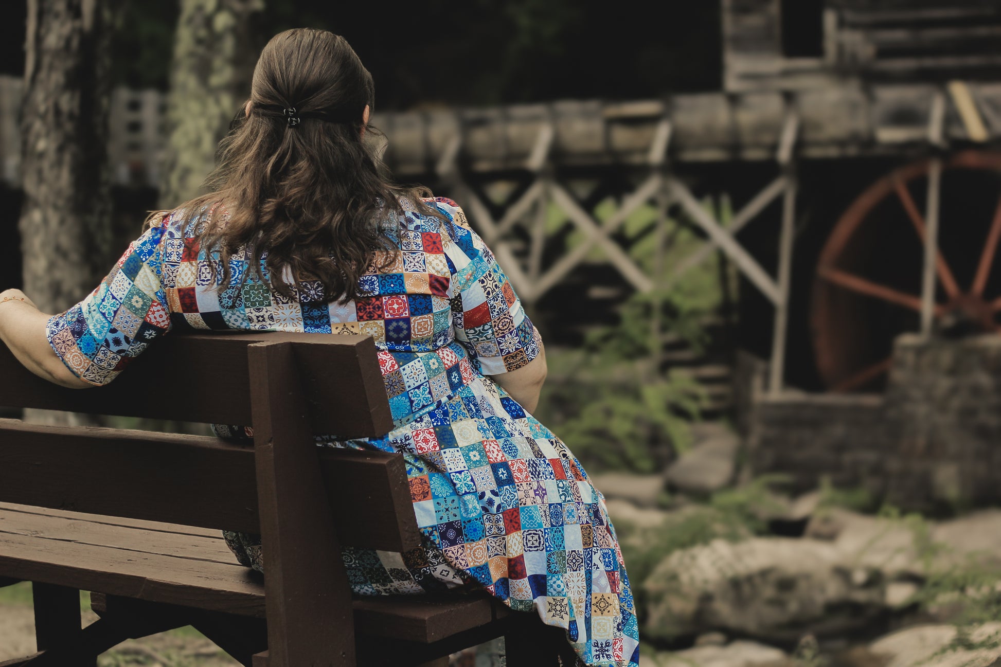 Person wearing a colorful patchwork modest nursing dress sitting on a wooden bench with a rustic background.