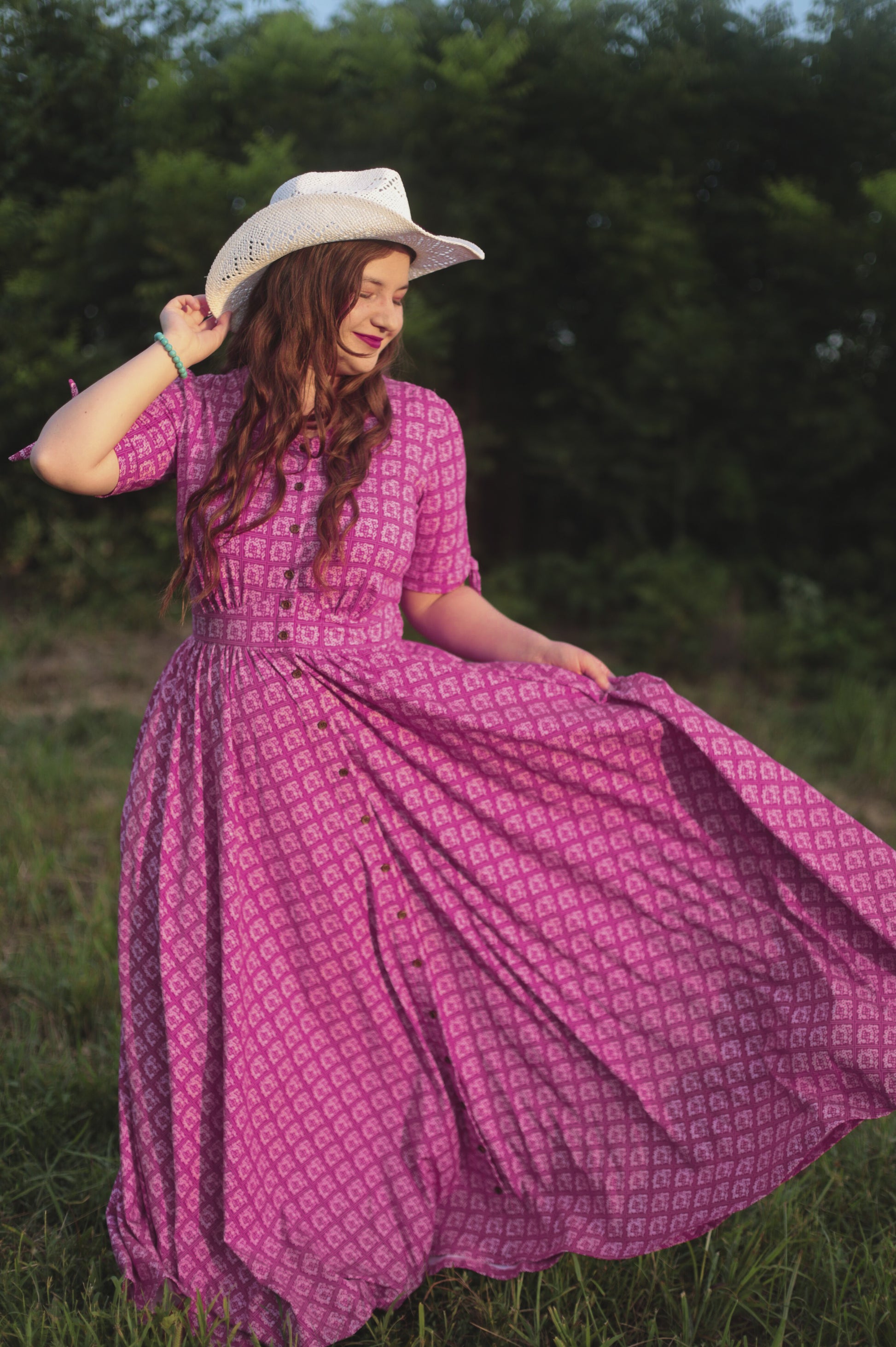 Woman in a pink modest nursing dress and cowboy hat standing in a grassy field.