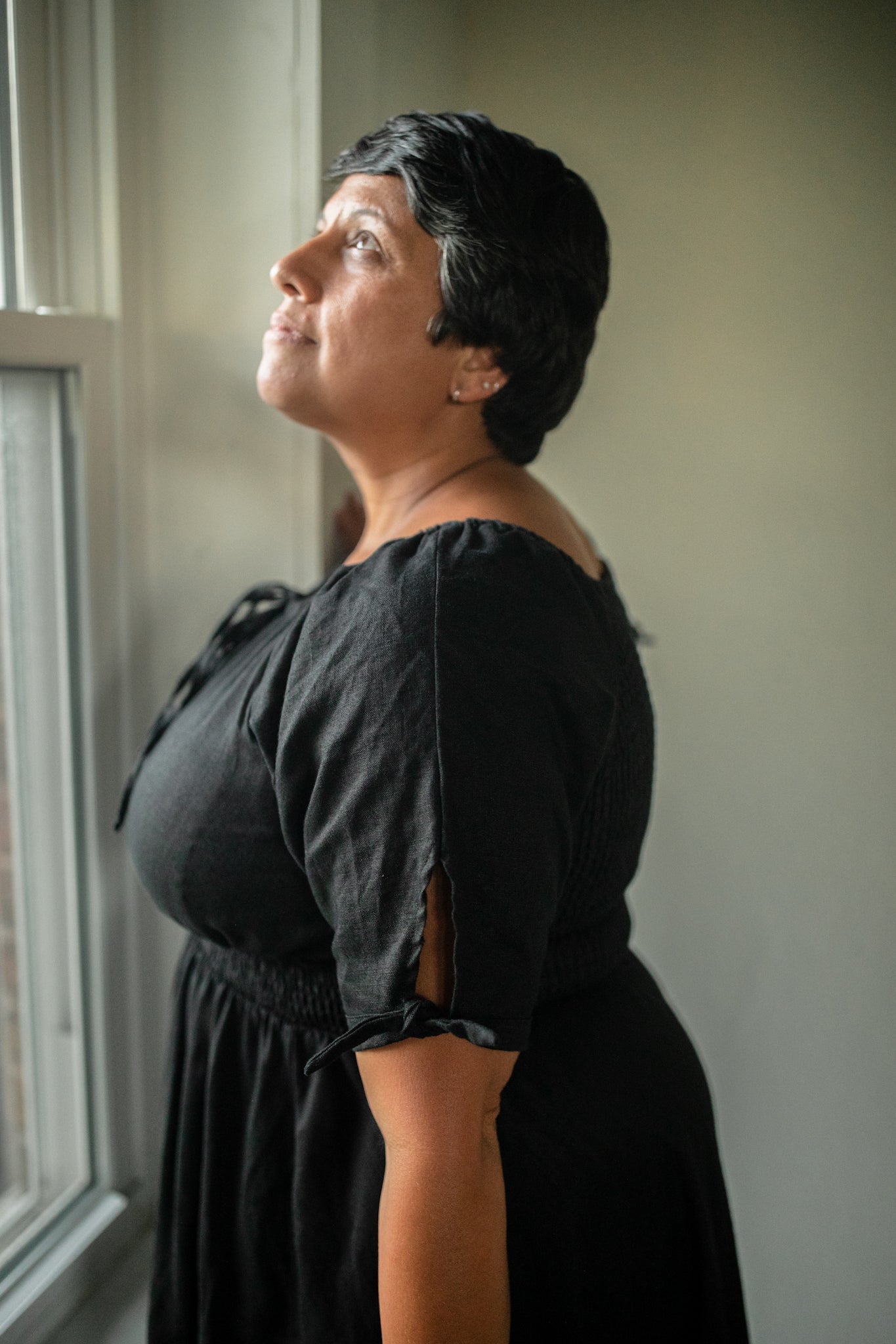 Woman in a black modest nursing dress standing by a window, looking upwards.