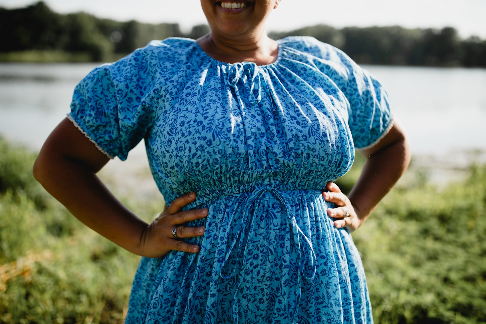 Woman wearing a blue modest nursing dress with a floral pattern standing outdoors by a body of water.