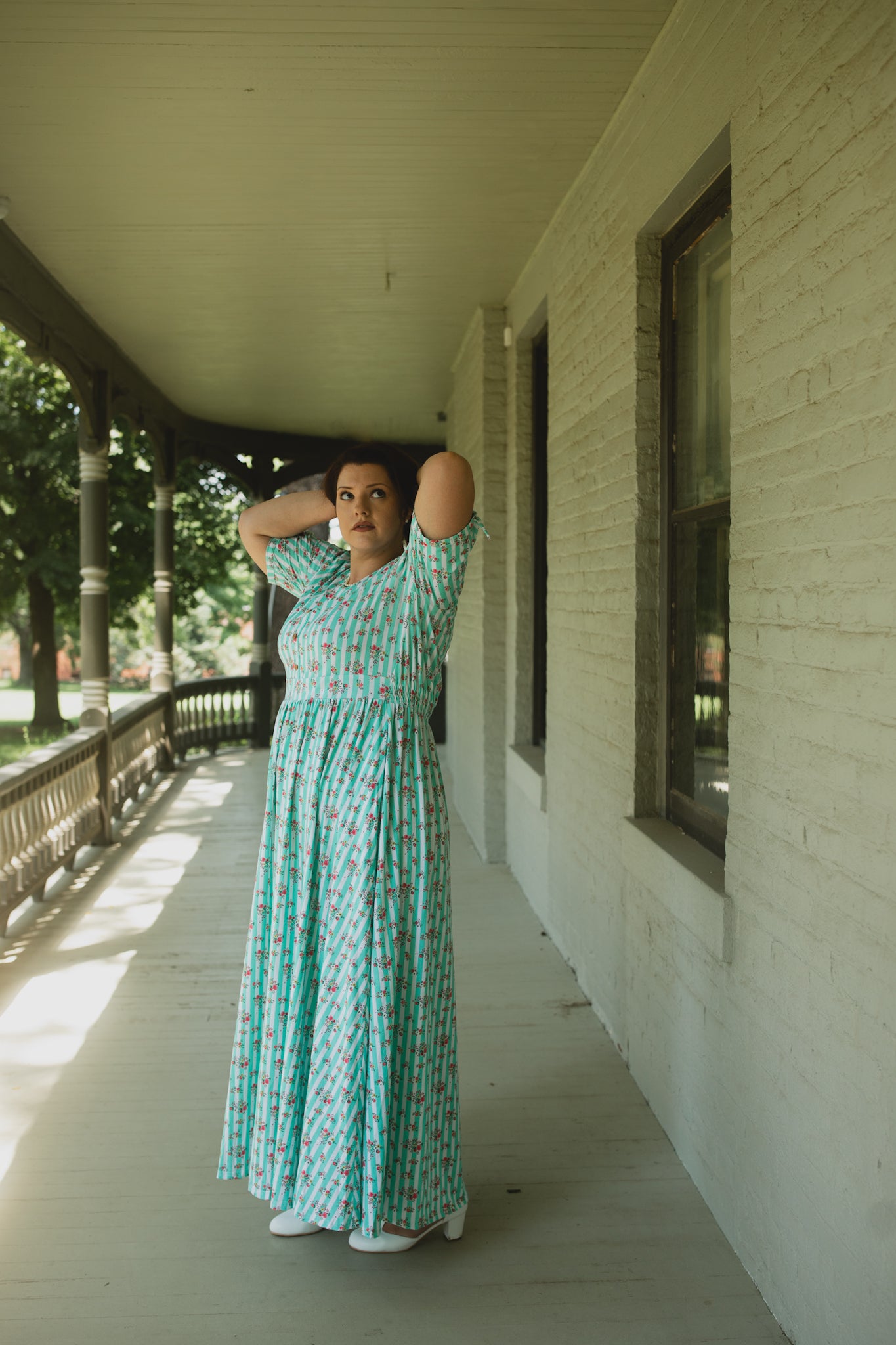 Woman in a green patterned modest dress standing on a porch.