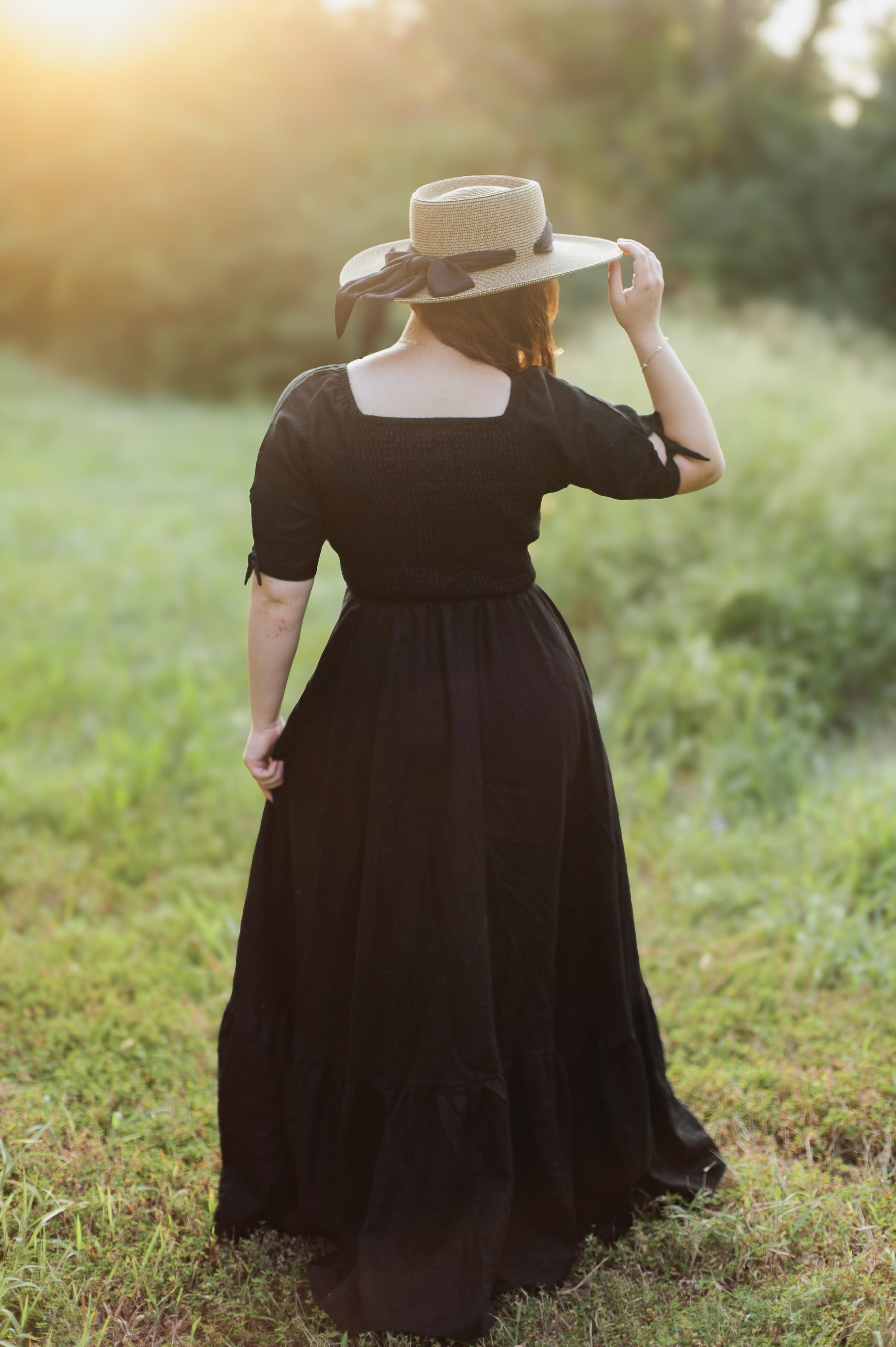 Woman in a black modest nursing dress and hat standing in a grassy field with a blurred background