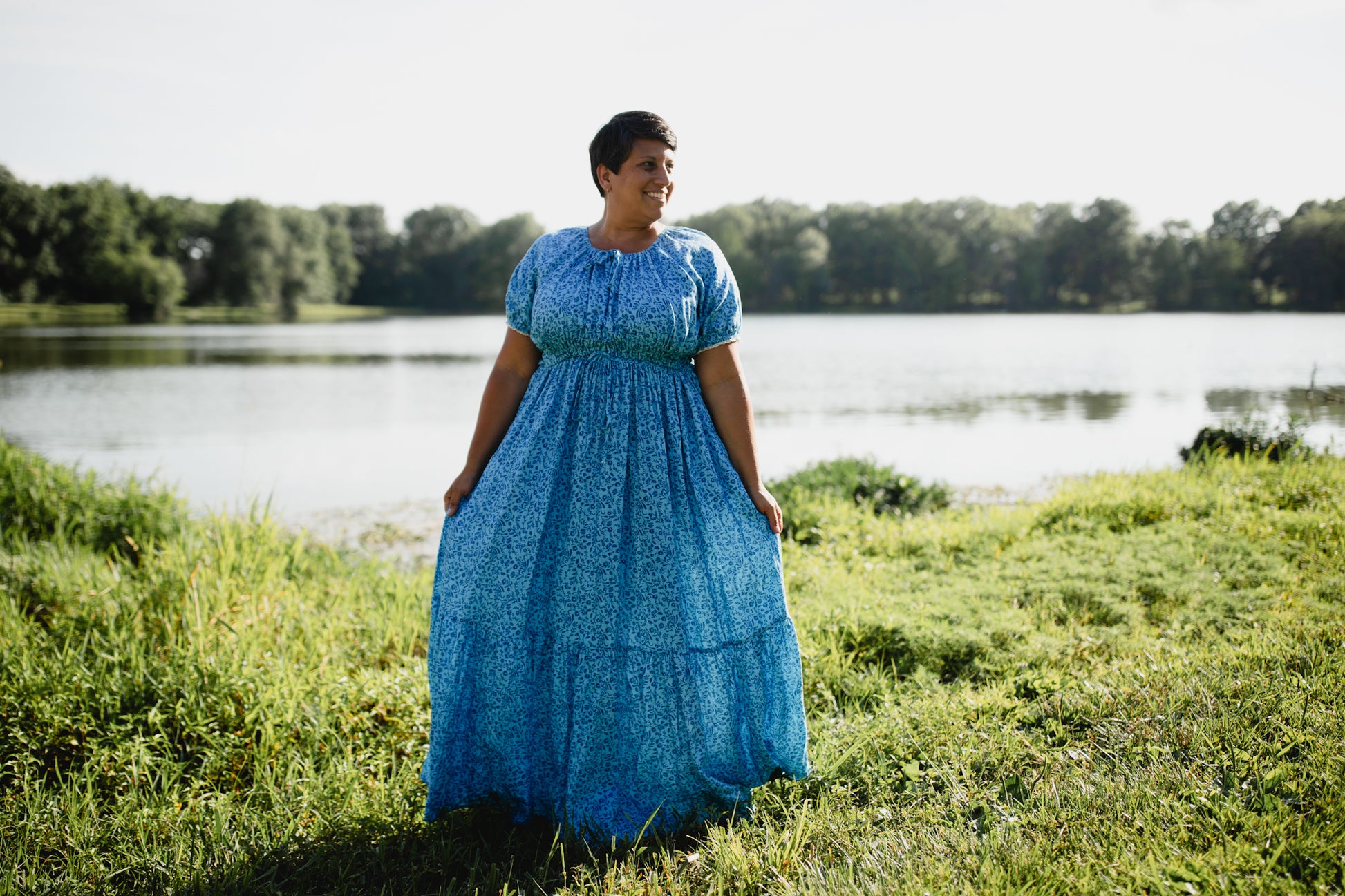 Woman in a blue modest nursing dress standing in a grassy area near a body of water.