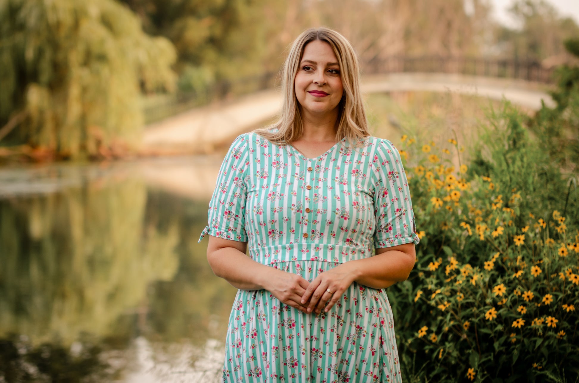 Woman in a floral modest nursing dress standing by a pond with greenery and flowers in the background
