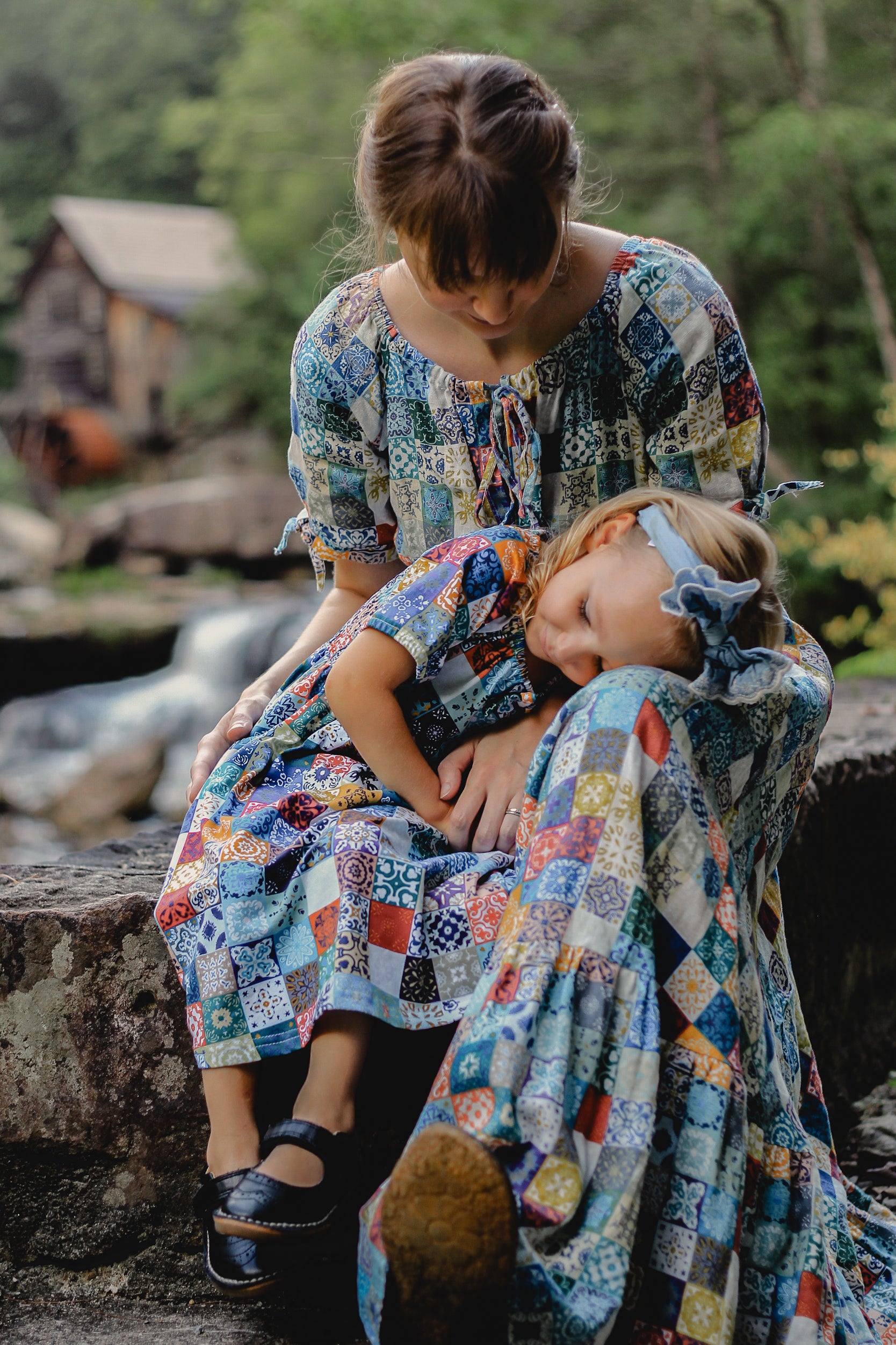 Mother and daughter in colorful patchwork modest dresses sitting outdoors by a stream.