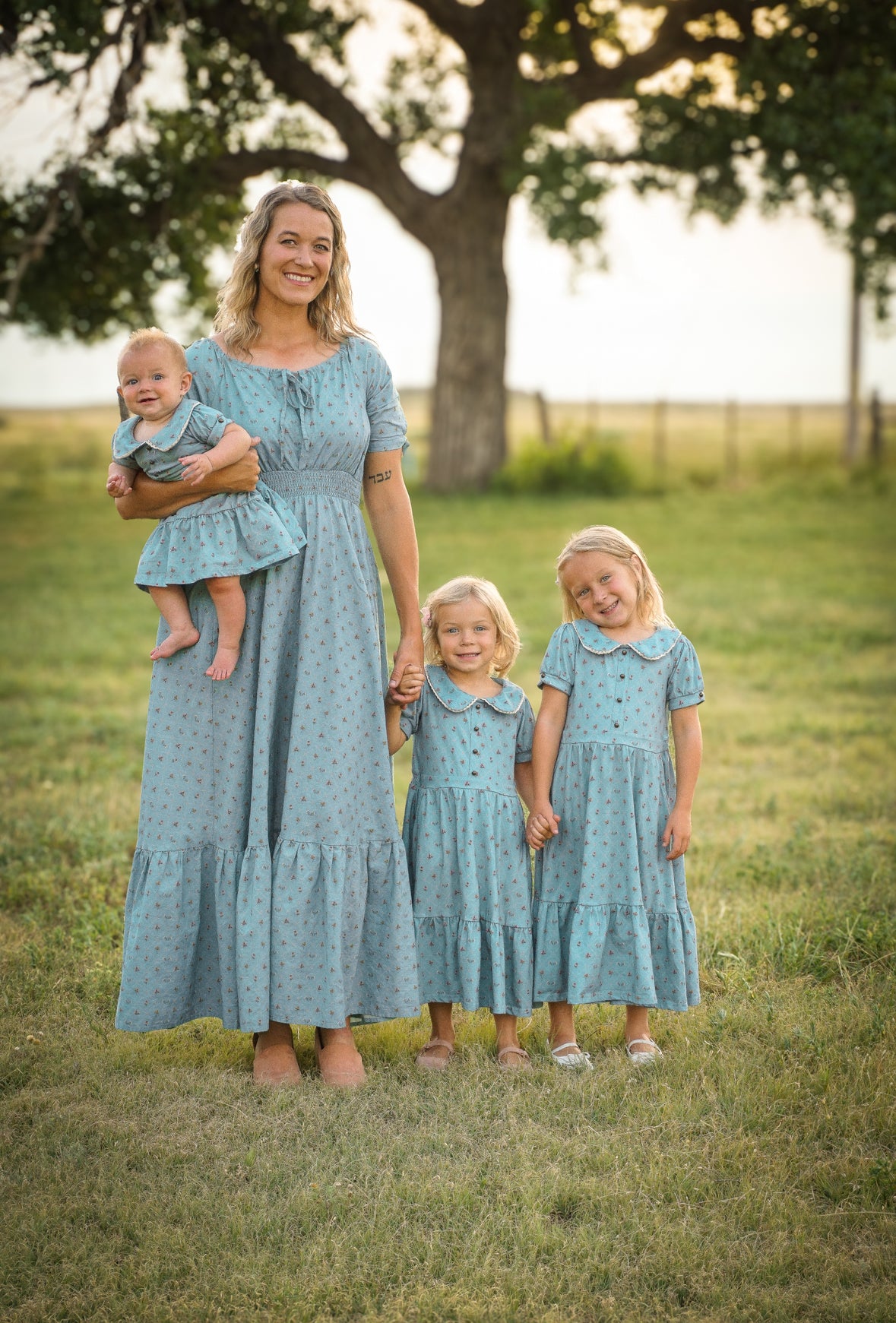Woman in a long blue modest nursing dress holding a baby, with two young girls in matching dresses standing on grass.
