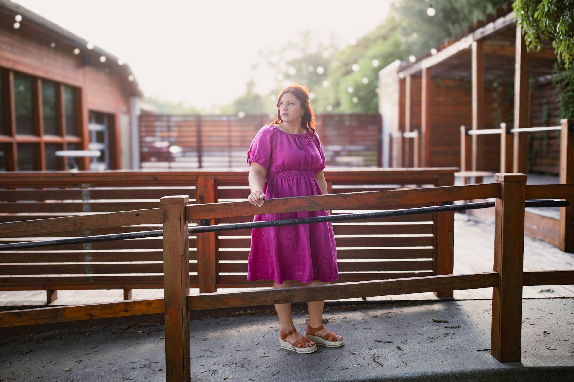 Woman in a pink modest nursing dress standing behind a wooden railing outdoors.