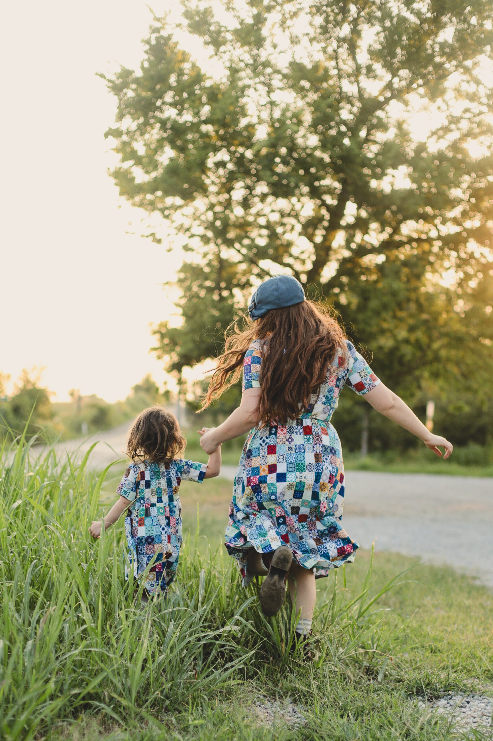 Mother and child in colorful modest dresses walking hand-in-hand through tall grass with trees in the background.