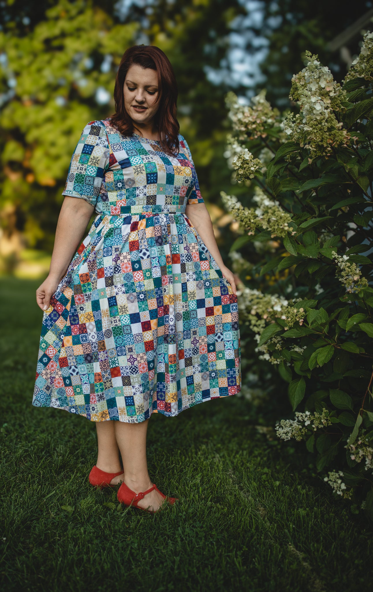Woman wearing a colorful patchwork modest nursing dress standing outdoors among greenery.