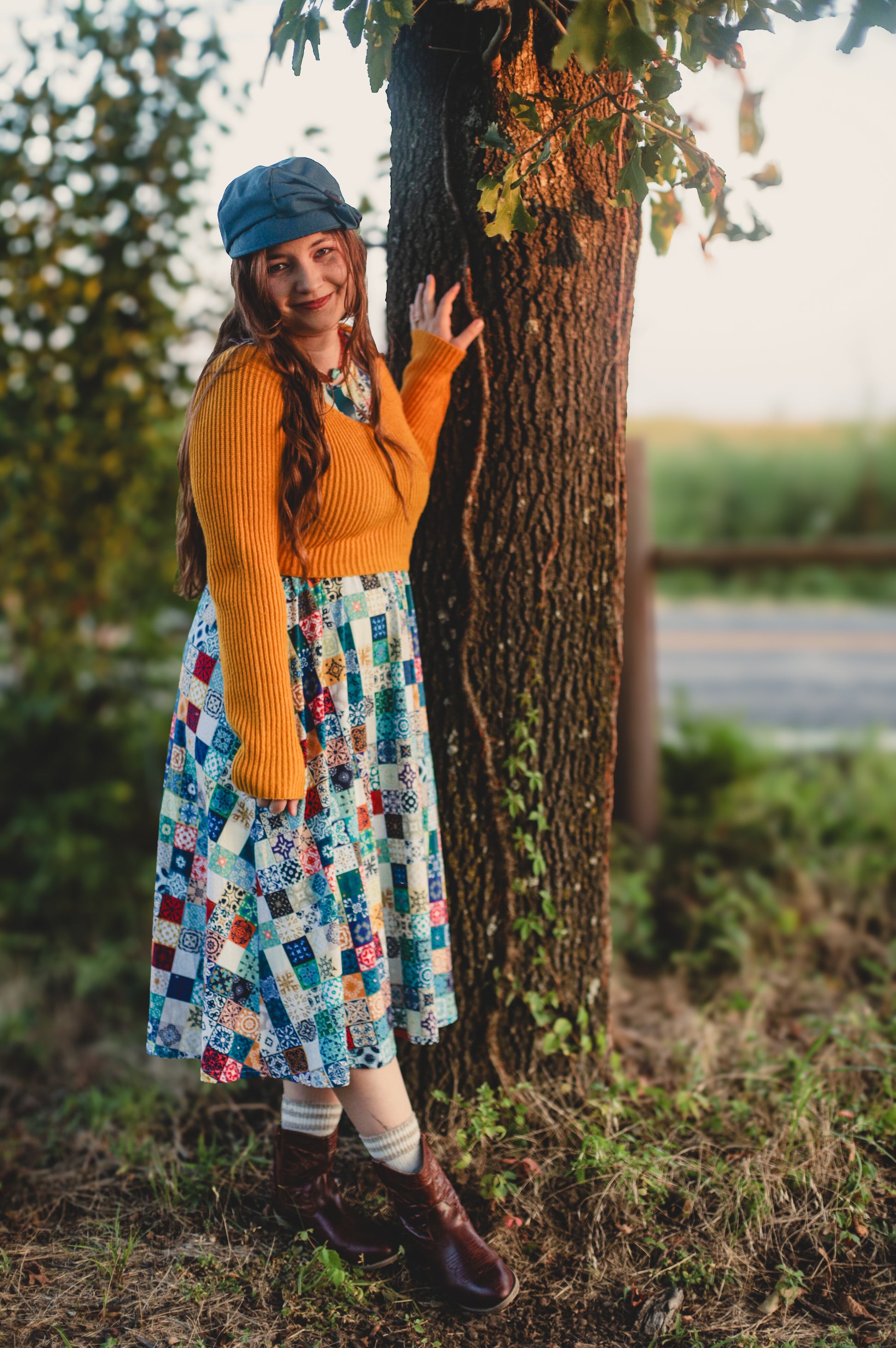 Woman in a colorful modest nursing dress and yellow sweater standing next to a tree outdoors.