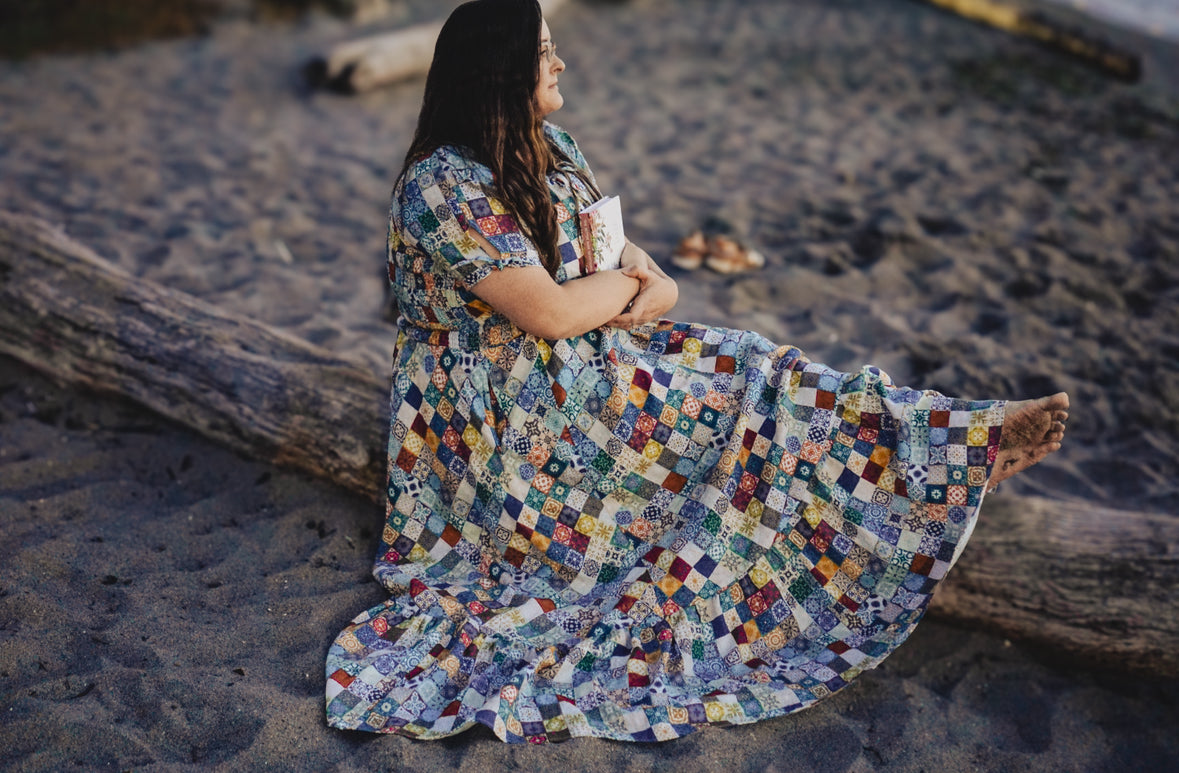 Woman in a colorful modest nursing dress sitting on a beach with a book