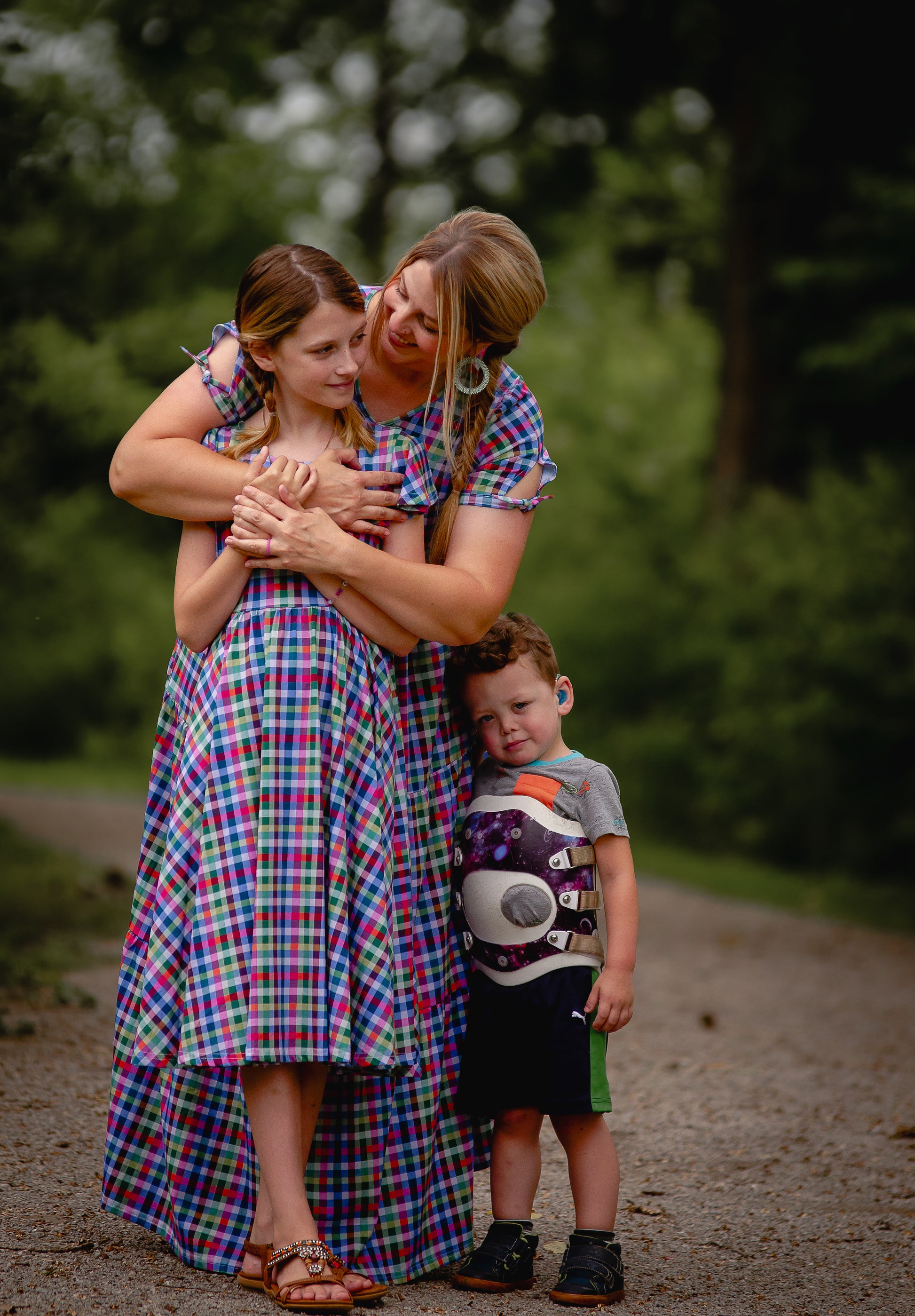 Mother and daughter hugging outdoors, wearing modest dresses, standing nearby, surrounded by greenery.