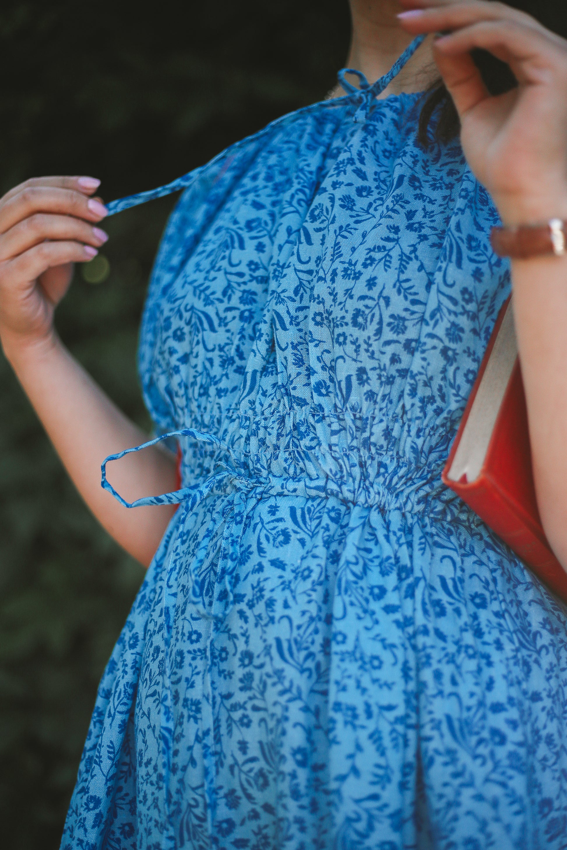 Woman wearing a blue floral modest nursing dress holding a red book against a blurred natural background