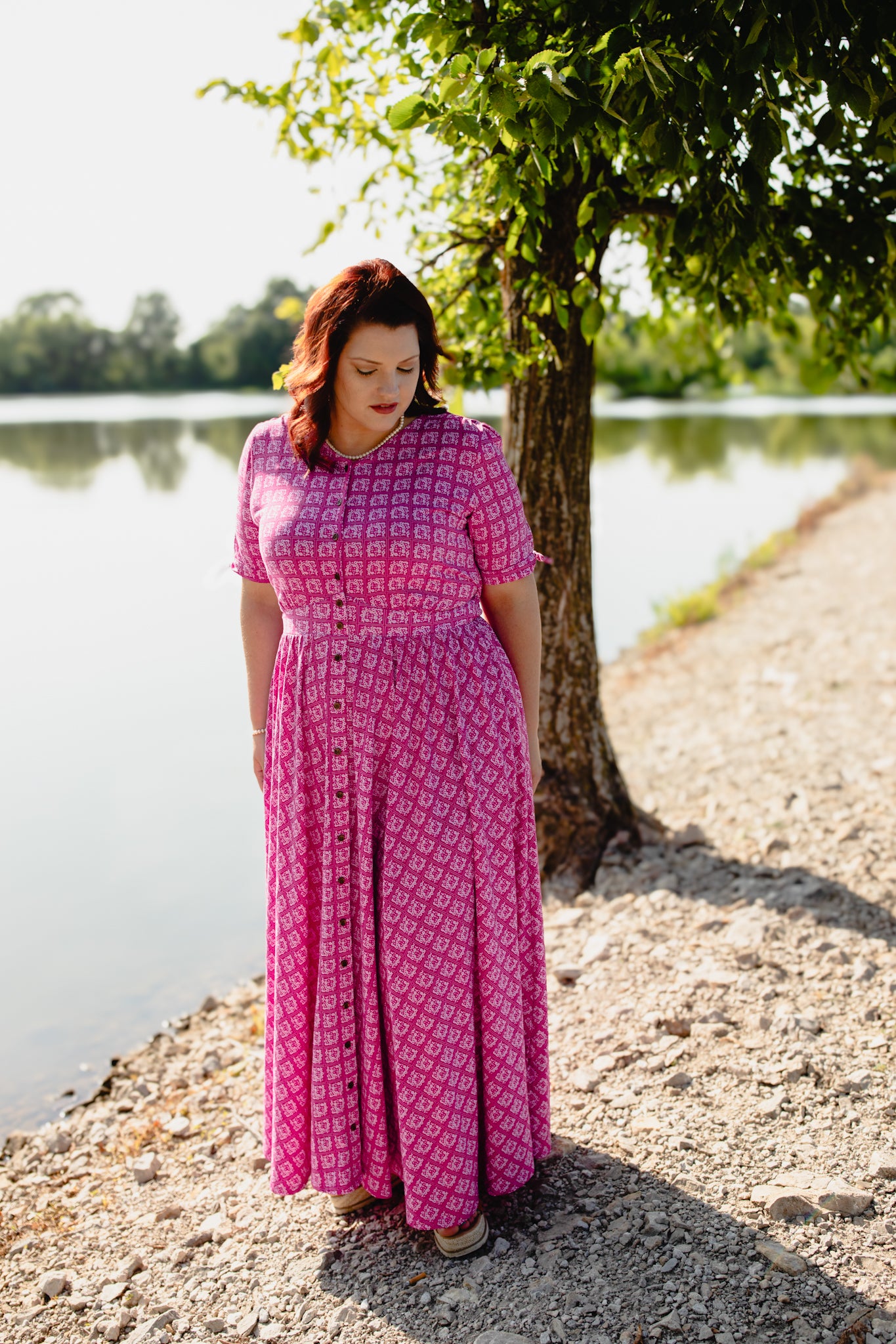 Woman in a pink modest nursing dress standing by a lake with trees in the background