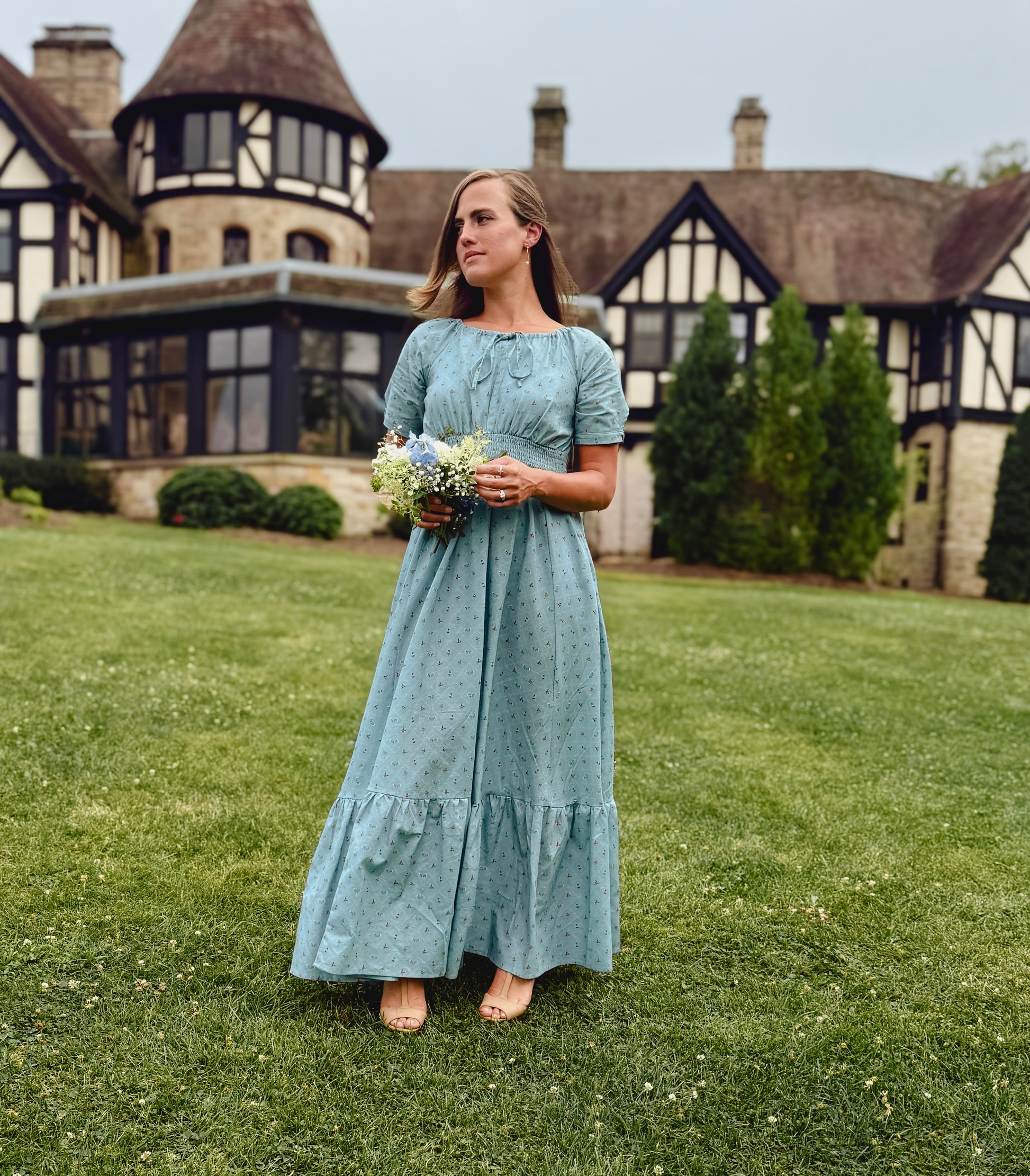 Woman in a blue modest nursing dress standing on grass in front of a large house