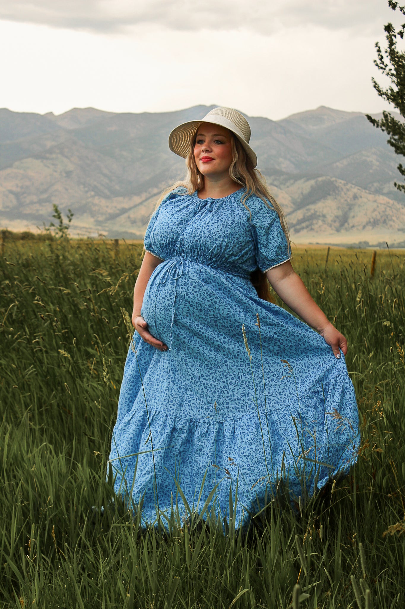 Woman in a blue modest nursing dress and hat standing in a grassy field with mountains in the background