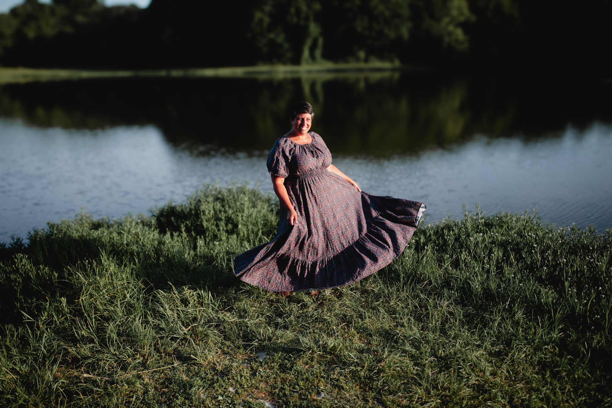 Woman in a dark modest nursing dress standing by a body of water with trees in the background
