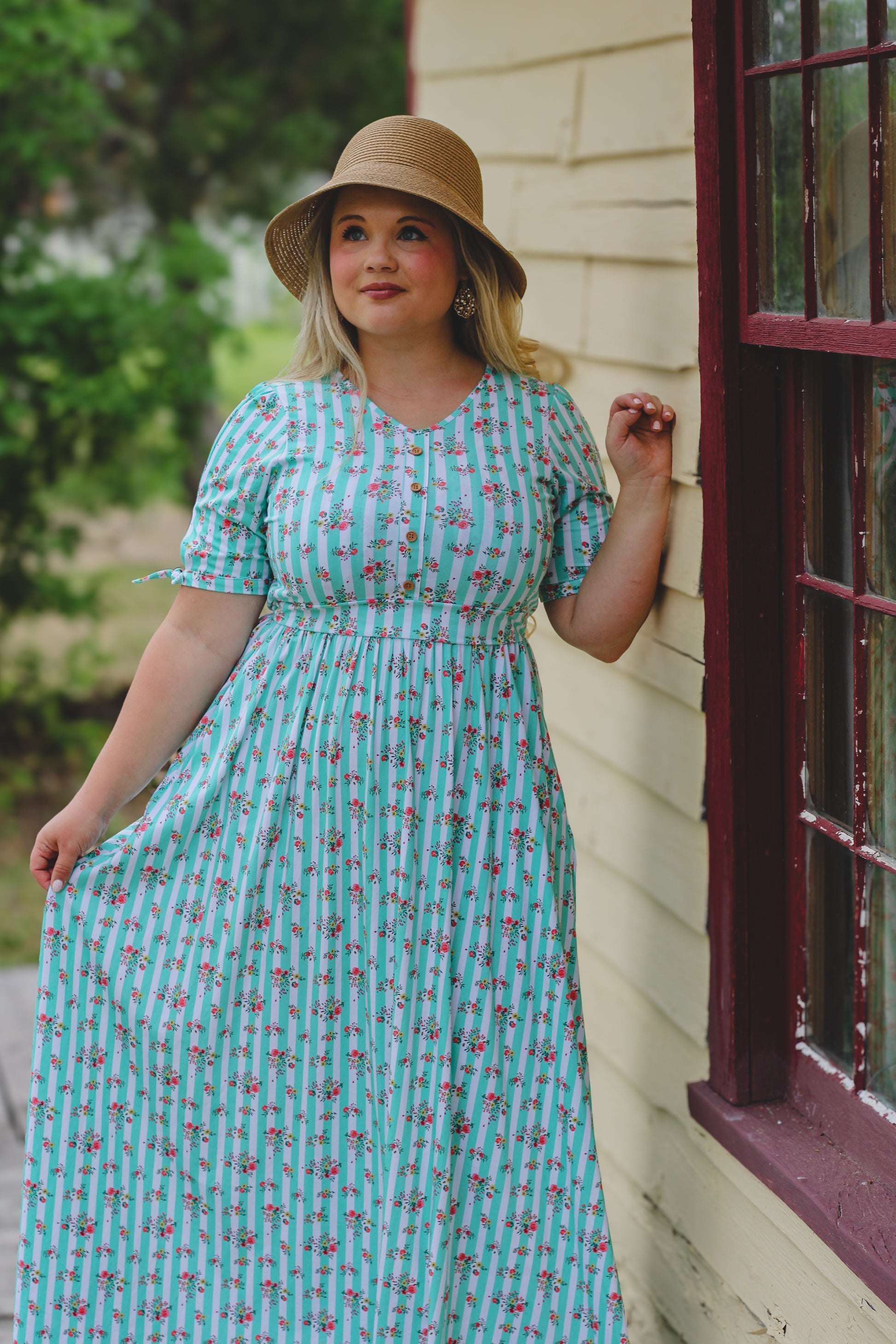 Woman in a teal floral modest nursing dress and hat standing against a house exterior.
