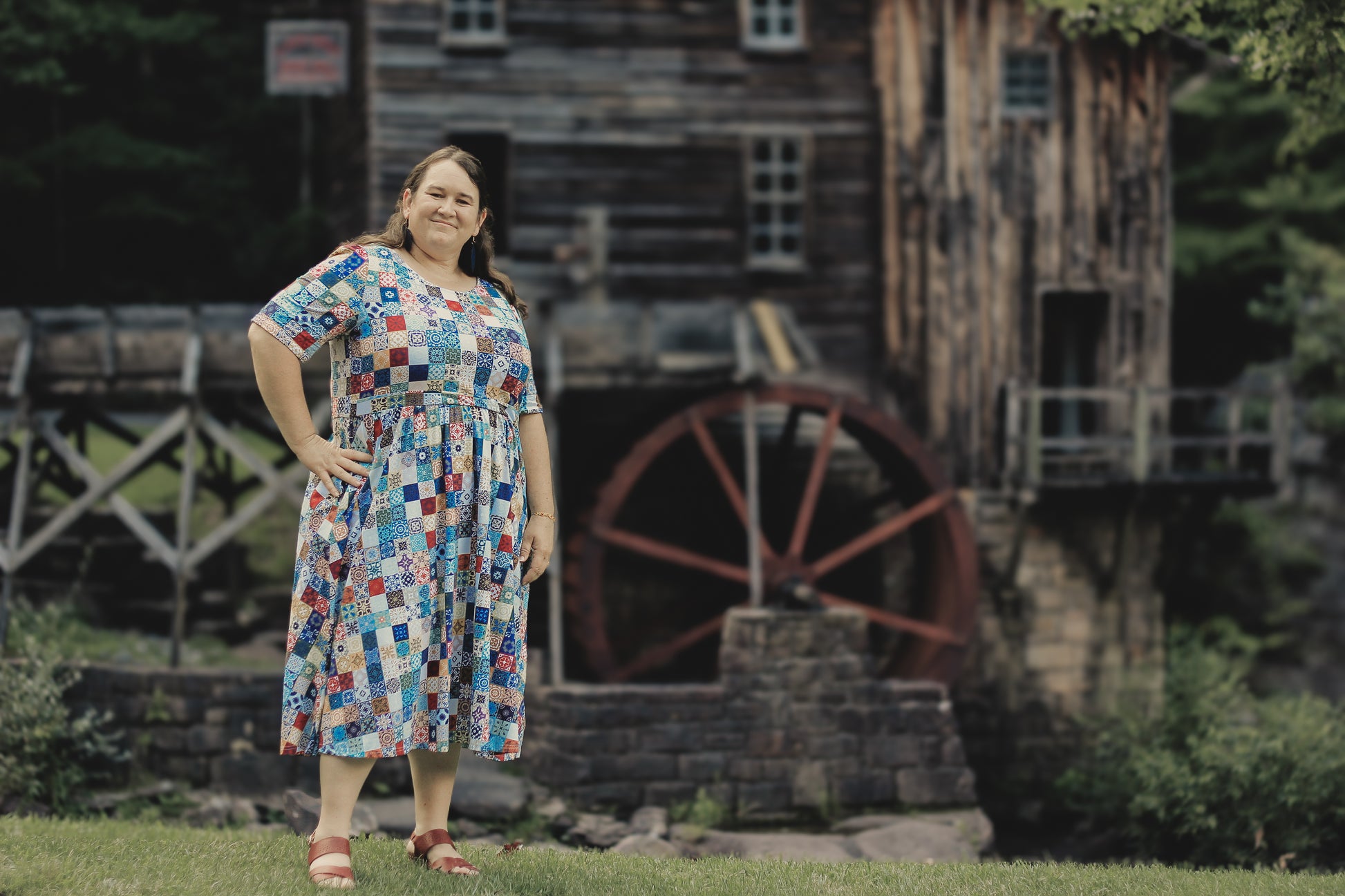 Woman in a colorful modest nursing dress standing in front of a watermill.