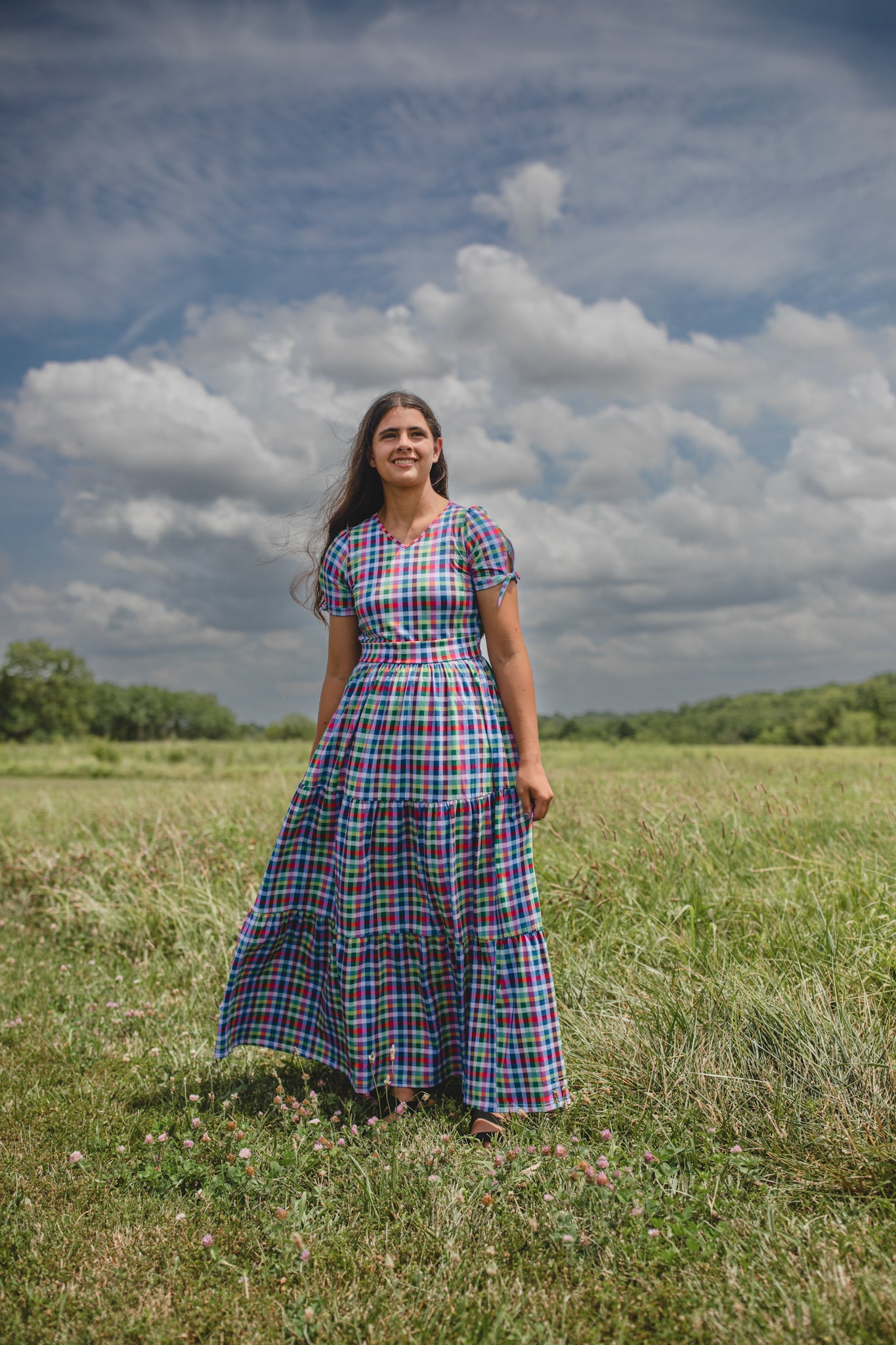 Woman in a plaid modest nursing dress standing in a field with a blue sky and clouds.