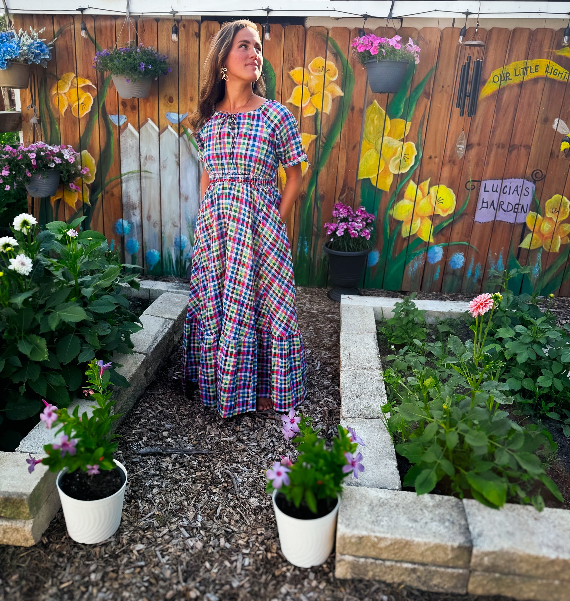 Woman in a plaid modest nursing dress standing in a garden with colorful flower pots and a wooden fence with floral designs.