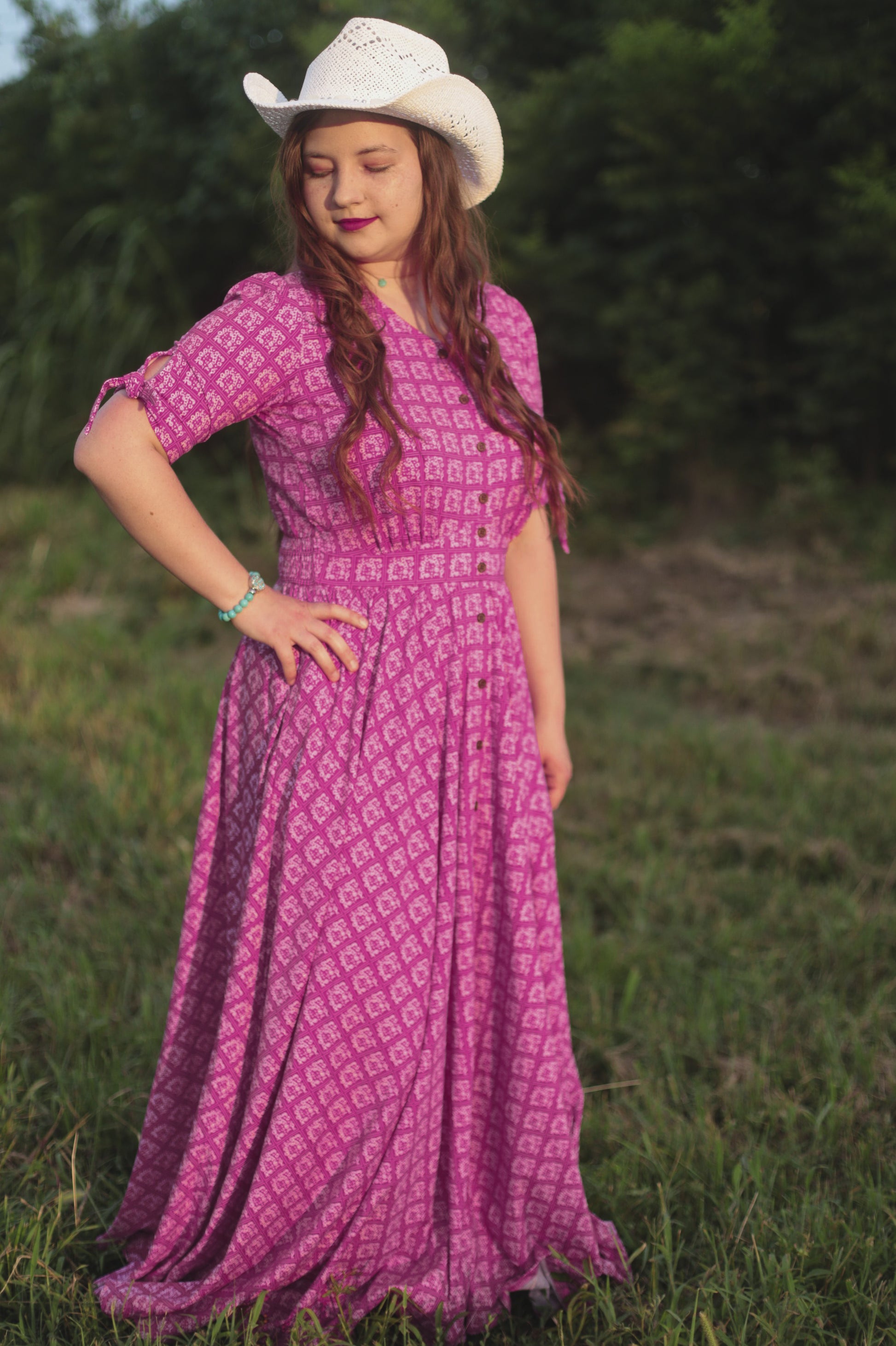 Woman in a purple modest nursing dress and white cowboy hat standing in a grassy field.