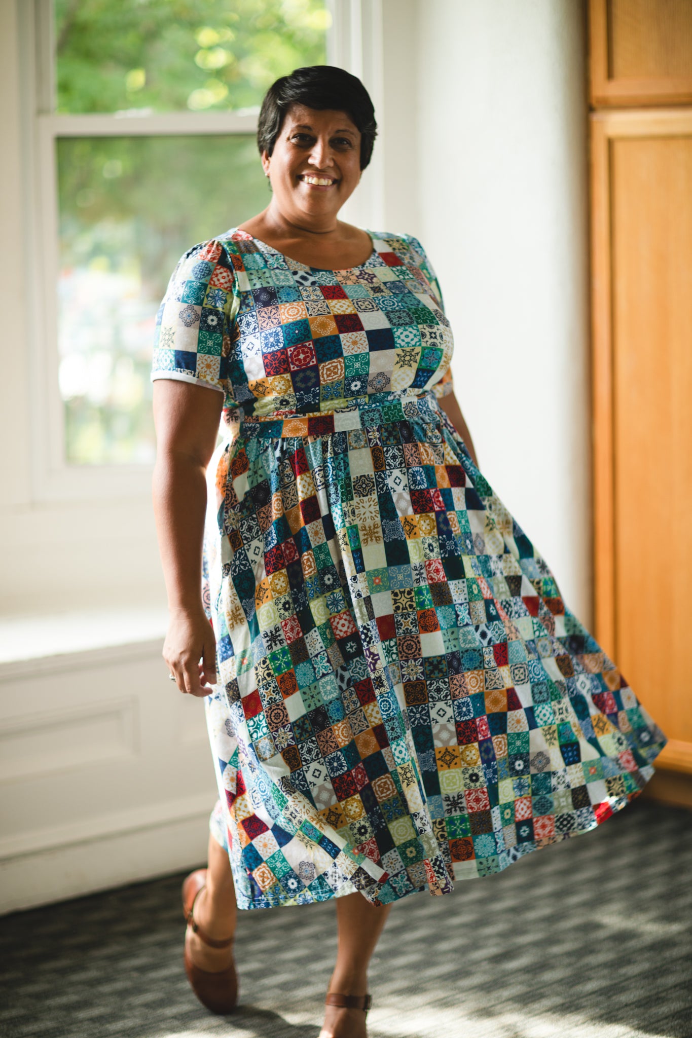 Woman wearing a colorful patchwork modest nursing dress standing indoors near a window.