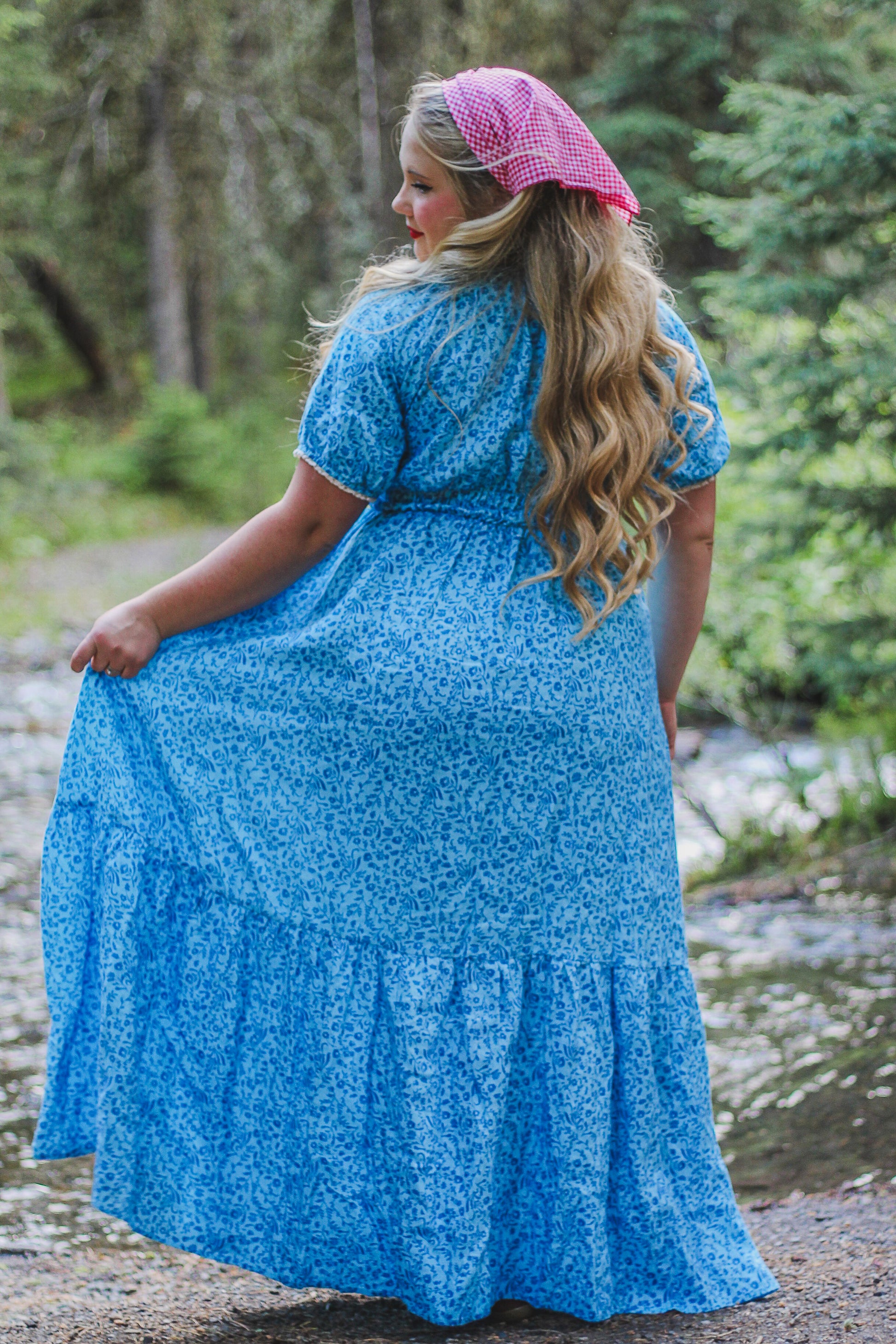 Woman in a blue modest nursing dress with a pink headscarf standing outdoors in a forest.
