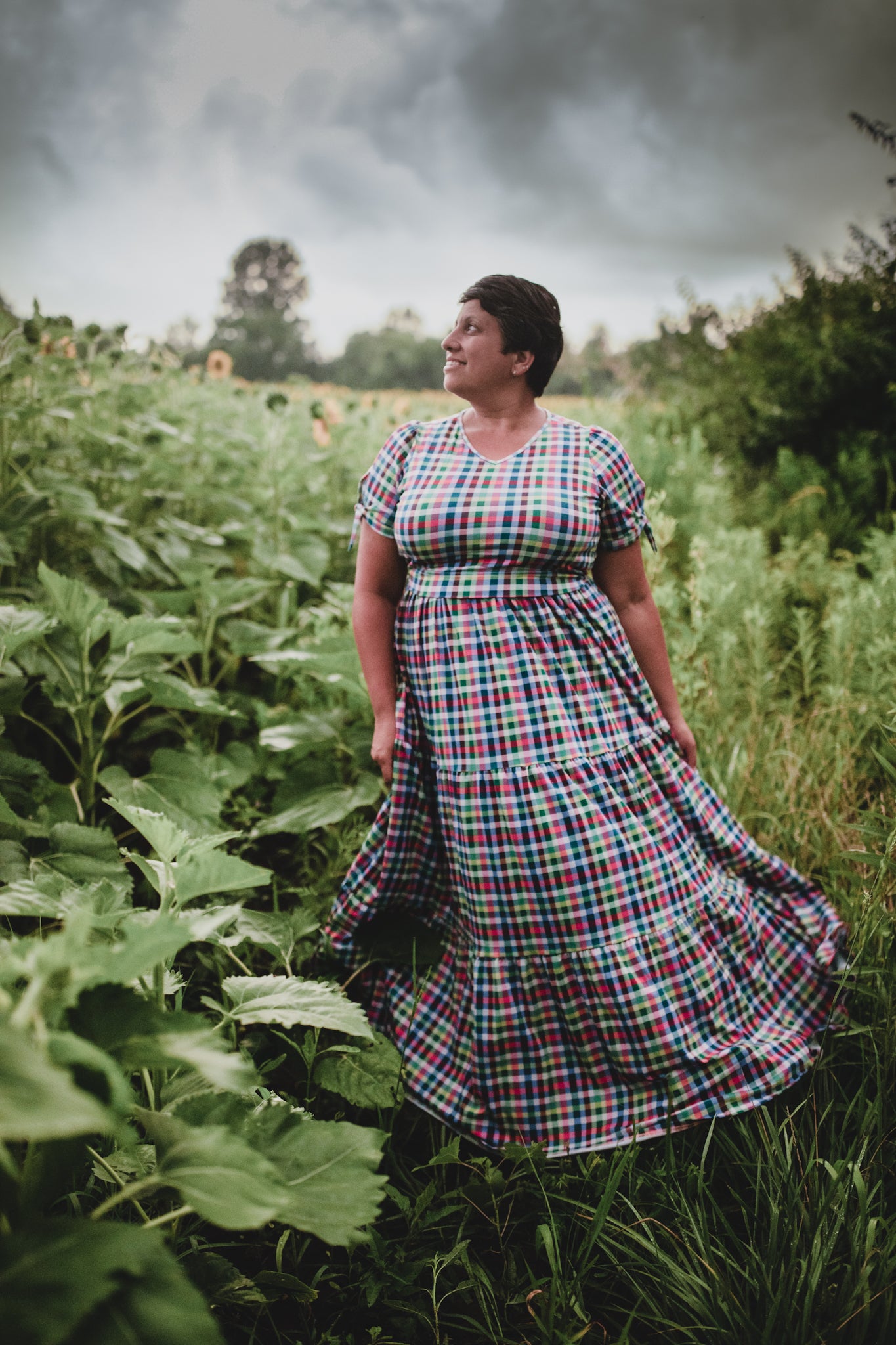 Woman in a plaid modest nursing dress standing in a field with stormy skies.